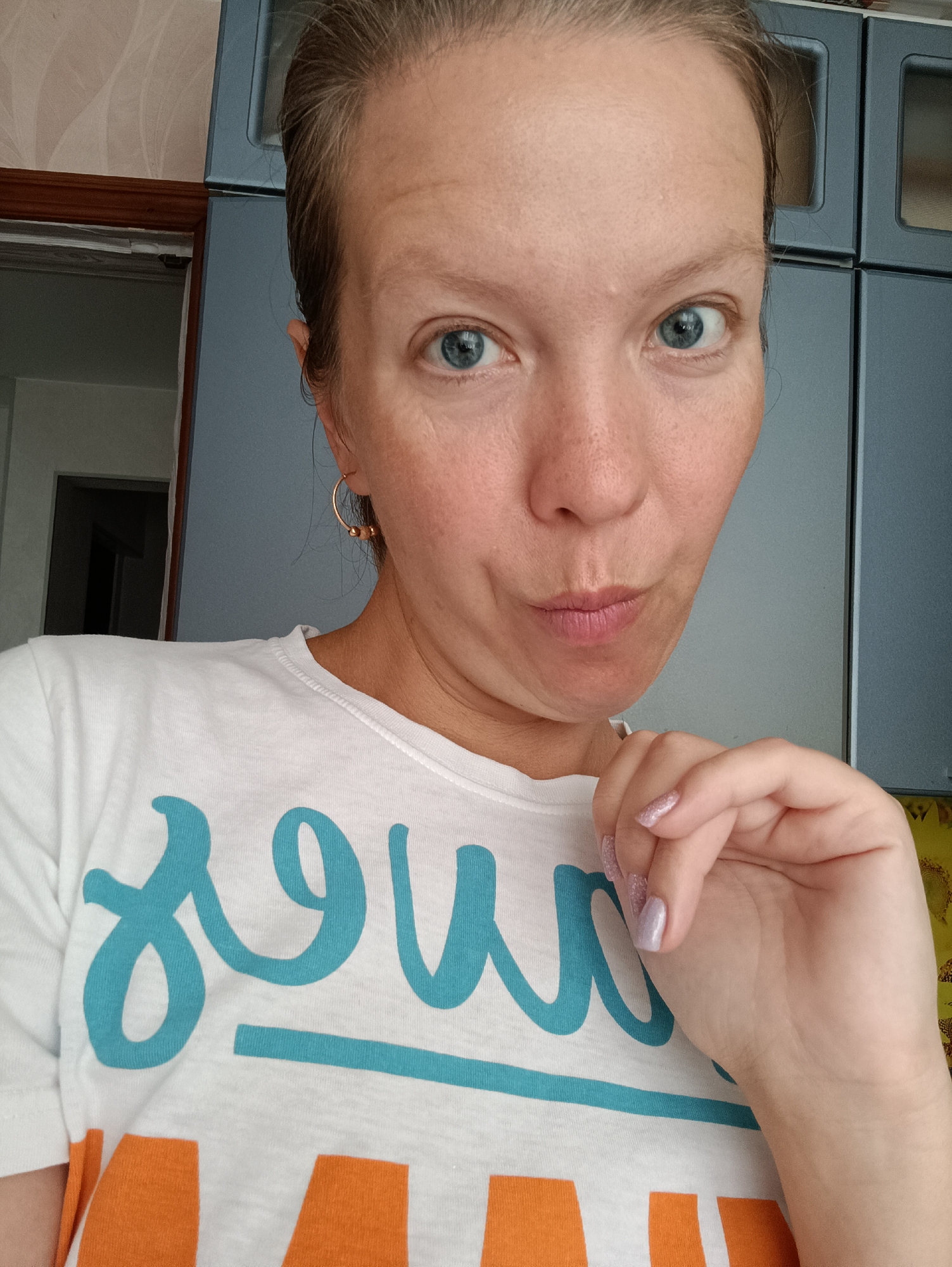 A single adult female is depicted in a close-up, chest-up indoor selfie originating from Chaikovsky, Russia. She has light skin with visible freckles, wide-set blue eyes, and light brown hair pulled back from her face. Her lips are slightly pursed, and she maintains direct eye contact with the camera. On her left ear, a gold-colored hoop earring with three small beads is visible. Her right hand, with fingers lightly curled and nails painted in a light, possibly glittery, polish, is positioned near her chin. She is wearing a white t-shirt featuring large, cursive-like blue lettering, partially obscured, and blocky orange lettering beneath it. The background to the viewer's left includes a light-colored wall with a faint pattern and a dark wooden door frame opening into a darker room. To the viewer's right, two sections of dark blue-gray cabinetry or panels, each with a recessed rectangular design, are visible.