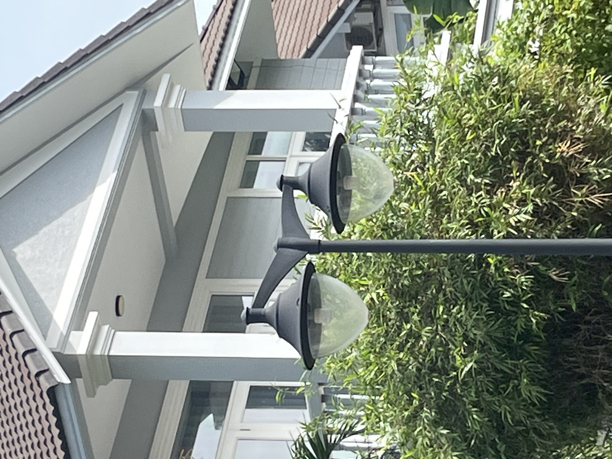 This is a medium shot of a street lamp with two globes in front of a house. The street lamp is made of dark metal and has a tall pole with two lamps attached at different heights. The lampshades are clear glass globes, and each contains a light bulb. The house is two stories and painted light gray with white trim. It has a tiled roof and several windows. Behind the street lamp and in front of the house is a dense growth of green bamboo. The scene appears to be in a residential area. There are no people visible in the image. The location is Phu Thuy Ward, Vietnam.