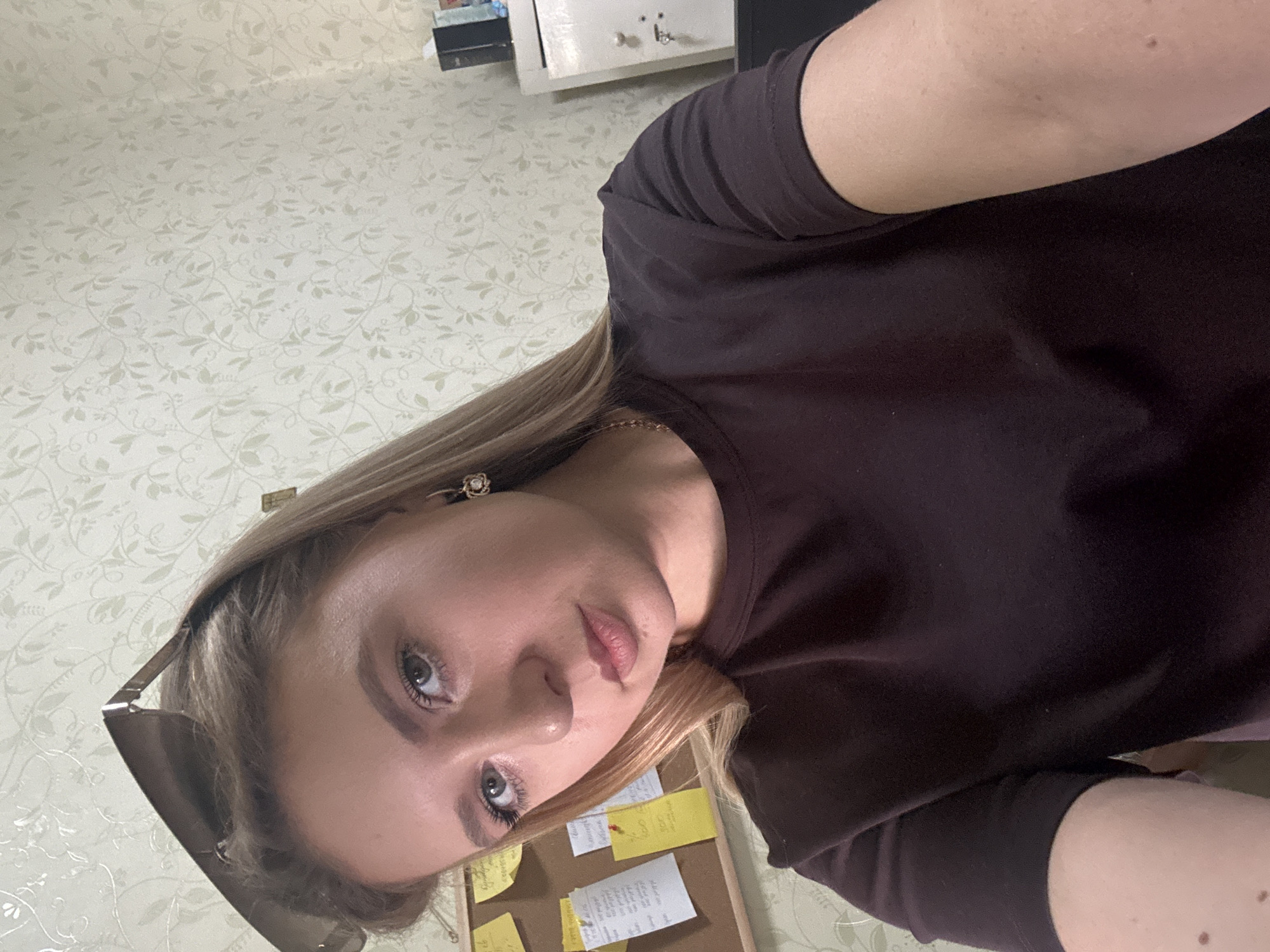 A young Caucasian woman with blonde hair, wearing sunglasses pushed back on her head and a dark t-shirt, is captured in a close-up selfie. She is positioned in front of a corkboard with various notes and papers attached to it, and a patterned wallpapered wall. To her right, a white cabinet with a metal door is visible, along with some black boxes on top of it. The woman is looking directly at the camera with a neutral expression. The overall scene appears to be an indoor setting, possibly an office or study space.