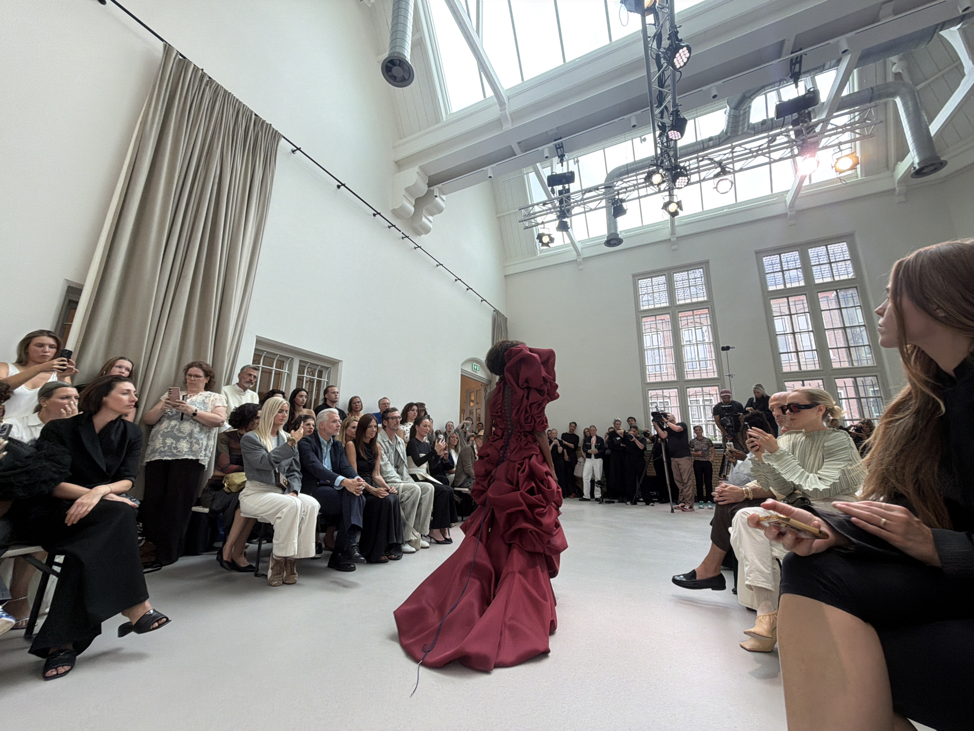 The image shows a fashion show in a large, high-ceilinged room with large windows. The room has white walls and a white floor. A model in a dramatic, voluminous red dress is walking down a runway. A crowd of people are seated in chairs on either side of the runway, watching the show and taking pictures with their phones. The people are dressed in a variety of fashionable clothing. The setting appears to be an indoor event space with industrial architectural elements visible, such as exposed ductwork and lighting rigs. The location is the Dom Church Tower in Utrecht, Netherlands.