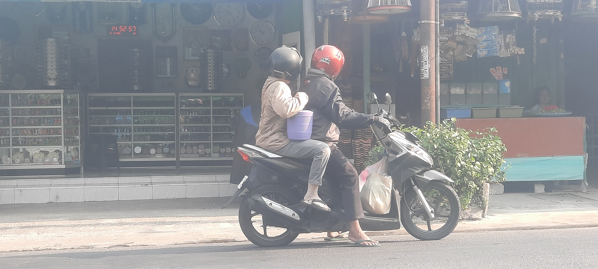 Two individuals are riding a motorcycle on a street in Selopuro, Indonesia. The person in front is driving and wears a red helmet, black jacket, and dark pants. The person behind is a passenger wearing a black helmet, a light brown jacket, and gray pants. They are carrying a light purple plastic bucket between them. A light-colored plastic bag is attached to the front of the motorcycle. The motorcycle is black and has a silver exhaust pipe.

In the background, a shop is visible with display windows filled with various items, possibly watches and other small goods. A digital clock on the shop displays "14 52 57". The shop facade has a wooden finish and a sign with text, partly obscured. To the right of the shop, a woman is partially visible behind a counter, with a platter of red items, possibly food, in front of her. Lush green foliage is growing in front of the shop. The scene appears to be during daylight.