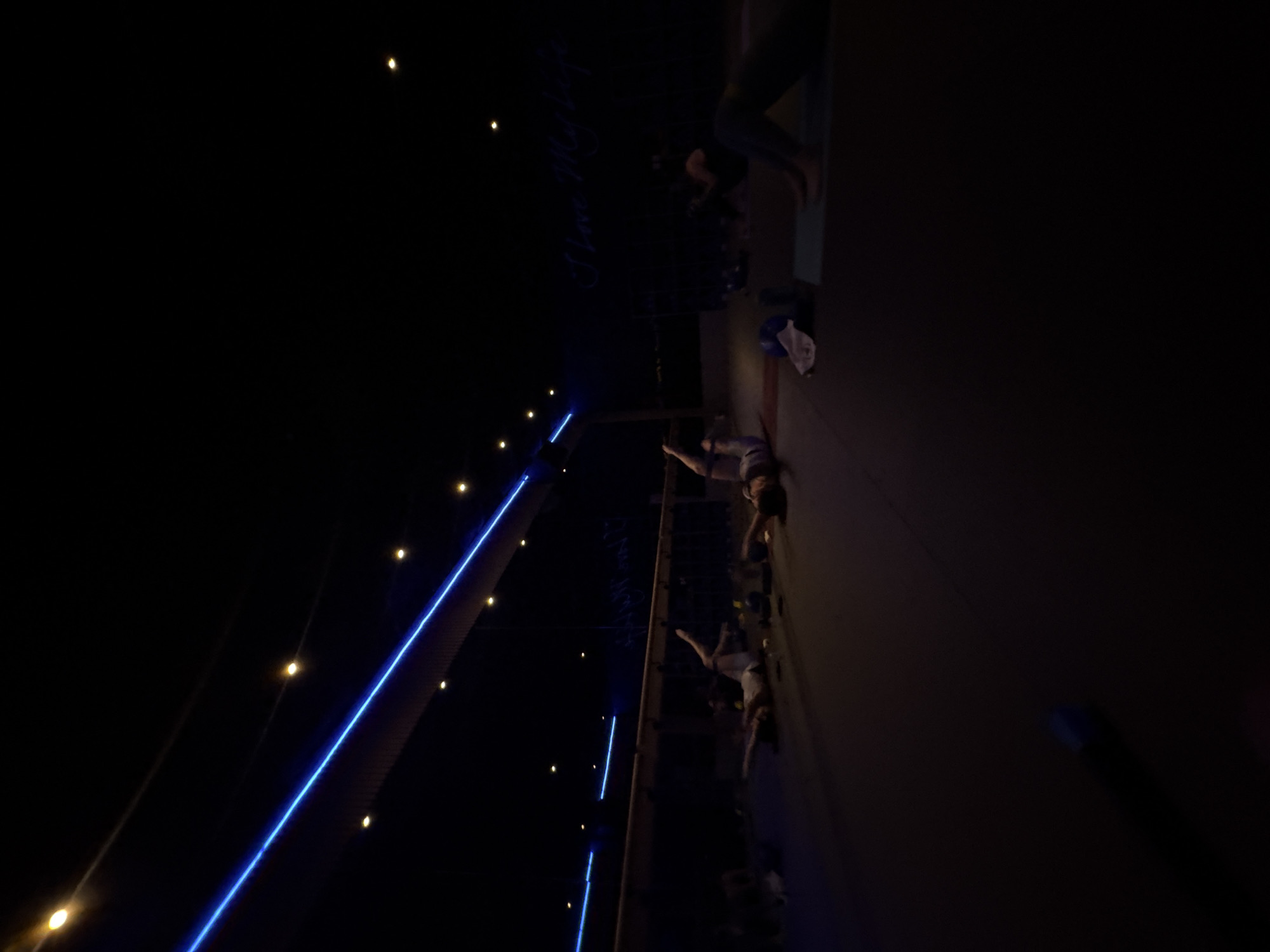 The image shows a nighttime scene in Mexico City, Mexico, with people engaging in a yoga session on a platform. String lights and blue neon lights illuminate the area. The participants appear to be performing yoga poses on mats. A yoga mat, a towel, and a water bottle are visible on the platform.