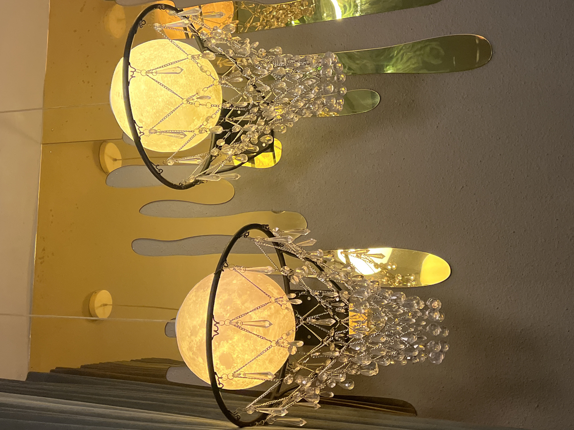 Two moon-shaped lamps are the primary focus, each surrounded by a black hoop and adorned with cascading crystals. The lamps are mounted on a wall with gold, cloud-like decorative panels. The background wall is a solid, neutral tone. The location is North Kuta, Indonesia.