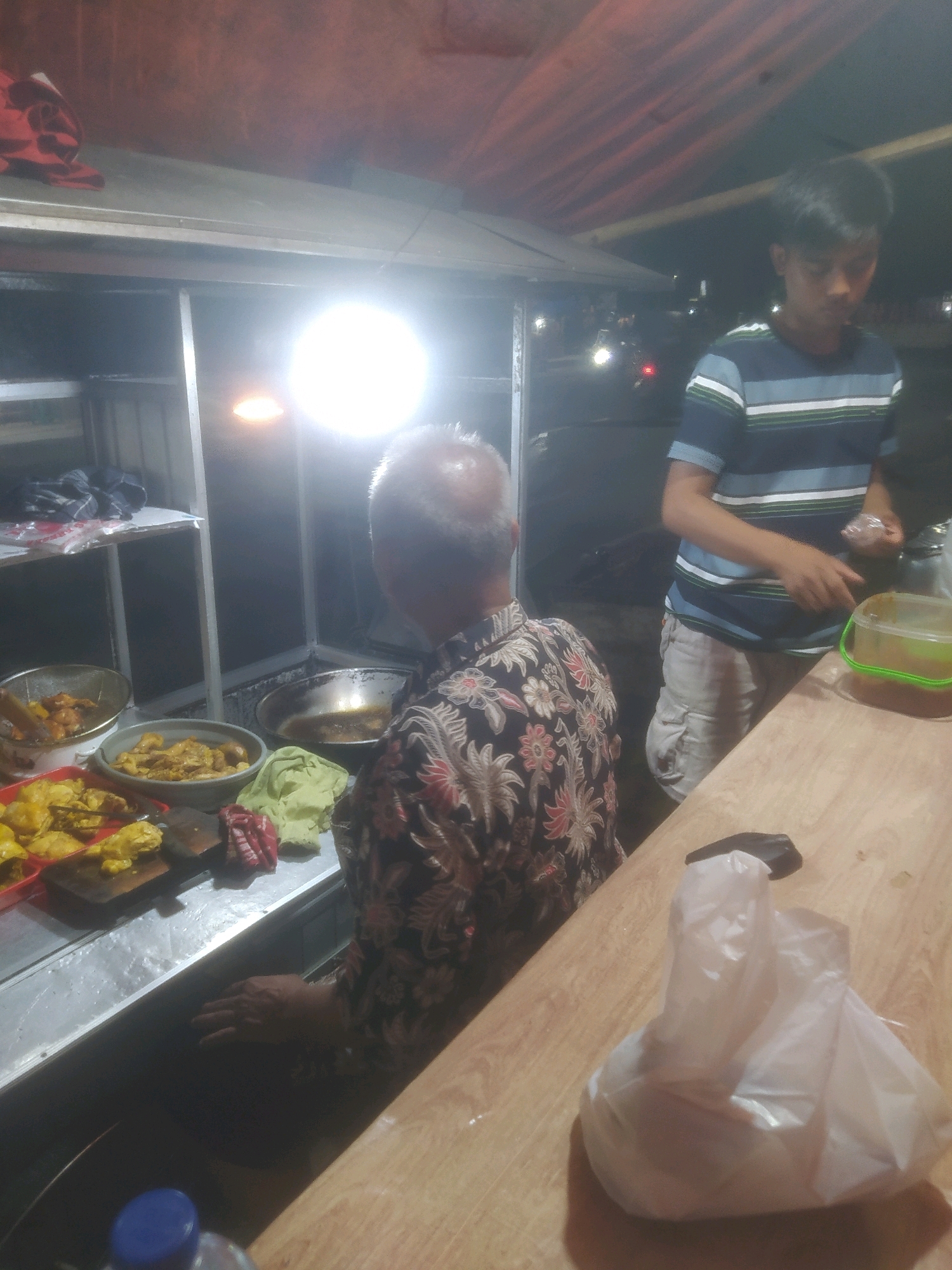 In Desa Karangbenda, Indonesia, a food stall is illuminated by a bright overhead light. A middle-aged man with graying hair, wearing a dark floral-patterned shirt, is positioned at the counter. He is facing away from the camera, looking towards a metal wok containing cooking oil, with a wooden cutting board and red and green cloths nearby. To his right, a younger man in a blue and white striped t-shirt and light-colored cargo shorts stands behind the counter. He holds a clear plastic bag in his right hand and appears to be preparing food. A plastic container with a green lid sits on the wooden surface in front of him. The stall is equipped with shelves holding various food items, including fried chicken pieces and other dishes in bowls. A clear plastic bag containing food is placed on the wooden counter in the foreground. The overall scene depicts a lively night market atmosphere.