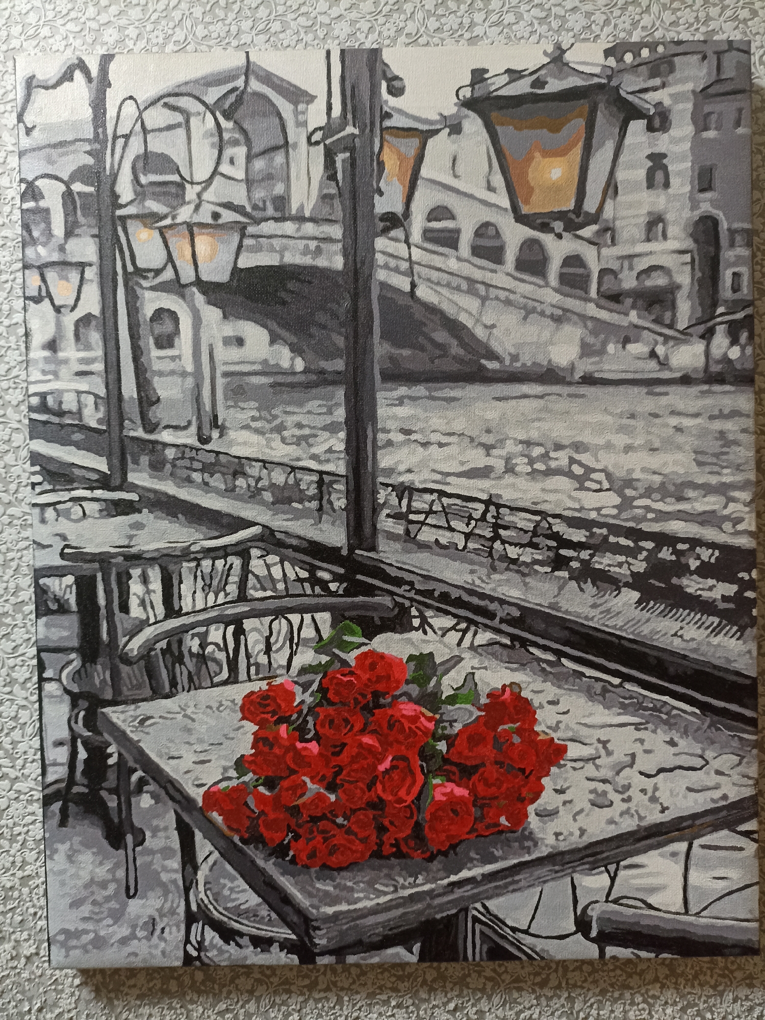 A painting depicts a picturesque scene, likely Venice, Italy, with a canal, a bridge, and ornate buildings in the background. The foreground features a table with a bouquet of red roses, set on a cobblestone or tiled surface that appears wet, suggesting recent rain or a misty atmosphere. Two chairs are partially visible, hinting at an outdoor cafe or seating area. Streetlights with warm, glowing lamps illuminate the scene, casting a romantic ambiance. The style of the painting is somewhat impressionistic, with broad strokes and a limited color palette of grays, blacks, whites, and a vibrant red for the roses. The overall mood is tranquil and evocative, perhaps suggesting a moment of quiet contemplation or romance. There is no visible text in the image. The provided location context "Чехов-2, Russia" does not align with the visual cues of the painting, which strongly suggest Venice.