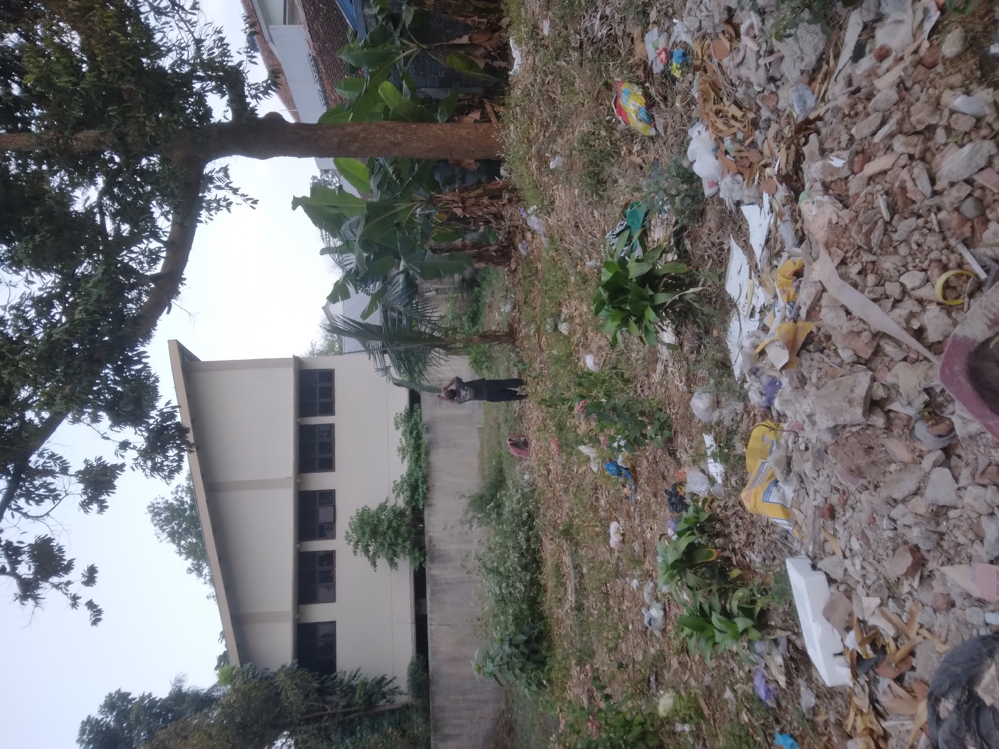 The image depicts a scene in Desa Pamijen, Kecamatan Sokaraja, Indonesia. In the foreground, a patch of ground is covered with debris and litter, including plastic bags, broken concrete, and other refuse. There are scattered plants and a pink backpack lying on the ground. In the middle ground, a person wearing a dark shirt and pants is standing, with their back to the camera, seemingly interacting with something near a tall plant. Behind the person, a concrete wall runs horizontally across the image. In the background, a cream-colored building with multiple dark windows is visible. To the right of the building, a large tree with dense foliage dominates the upper right portion of the image, with banana trees and other vegetation situated in front of it. A tiled roof of a building is also visible to the far right. The sky is overcast and appears to be either early morning or late afternoon.