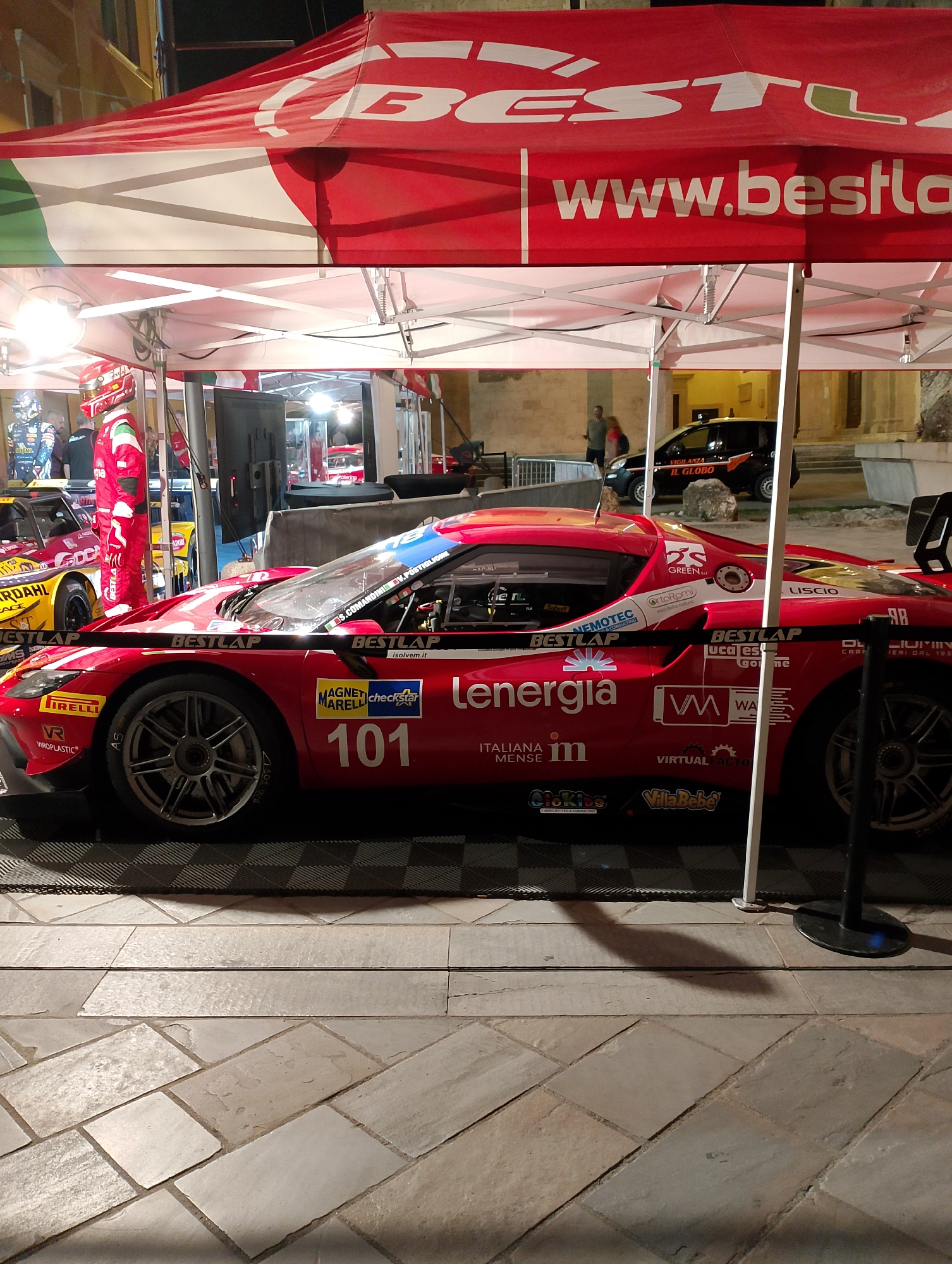 The image depicts a red racing car with the number 101 prominently displayed, parked under a red and white striped canopy. The car is adorned with various sponsor logos, including "BESTLAP," "LENERGIA," "MAGNARELLI," and "CHECKSTAR." A person in a red racing suit stands to the left of the car, facing away from the camera. Behind the car, another racing car, predominantly yellow, is visible. In the background, a dark-colored vehicle with "IL GLOBO" written on its side is parked. The scene is set on a cobblestone pavement, suggesting an outdoor urban environment. The presence of the racing cars and canopy implies an event related to motorsports. The provided context identifies the city as Rieti, Italy.