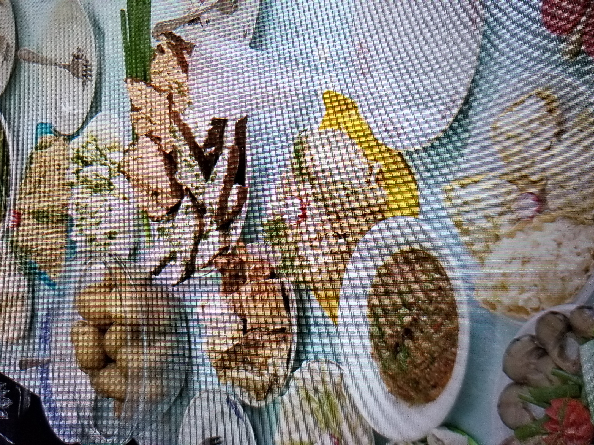 An overhead shot depicts a table laden with a spread of food items, seemingly in a domestic setting, possibly a home in Tiraspol, Moldova. The dishes include a glass bowl filled with small, whole potatoes, a plate of what appears to be bread with toppings, and a mound of crumbled white food, possibly rice or a salad, garnished with radishes. Another dish contains a reddish-brown paste or dip, and a plate of what looks like pickled mushrooms or olives, accompanied by sliced tomatoes and greens. The table is covered with a light blue cloth, and various plates, some with decorative rims, are visible. The lighting suggests an indoor environment.