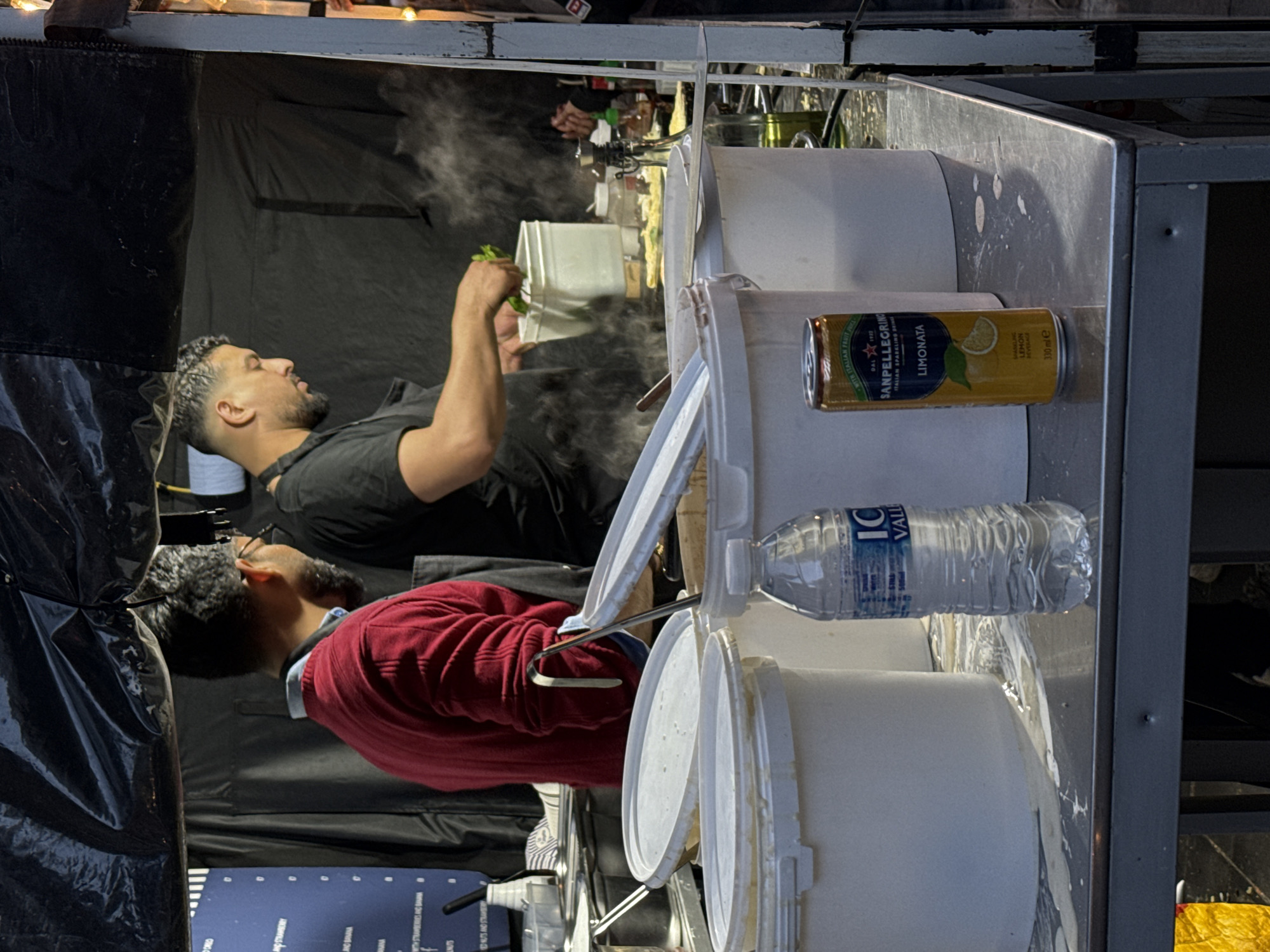 Two men are working inside what appears to be a food stall. One man, wearing a black t-shirt, is holding a white container with leafy greens, possibly preparing food. The other man, in a red sweater, appears to be stirring something in a large container. There are several white buckets and containers, along with a bottle of "IC Valley" water and a can of "San Pellegrino Limonata" on a metal counter, indicating that drinks are available. The location is not immediately clear in the city of London, United Kingdom.