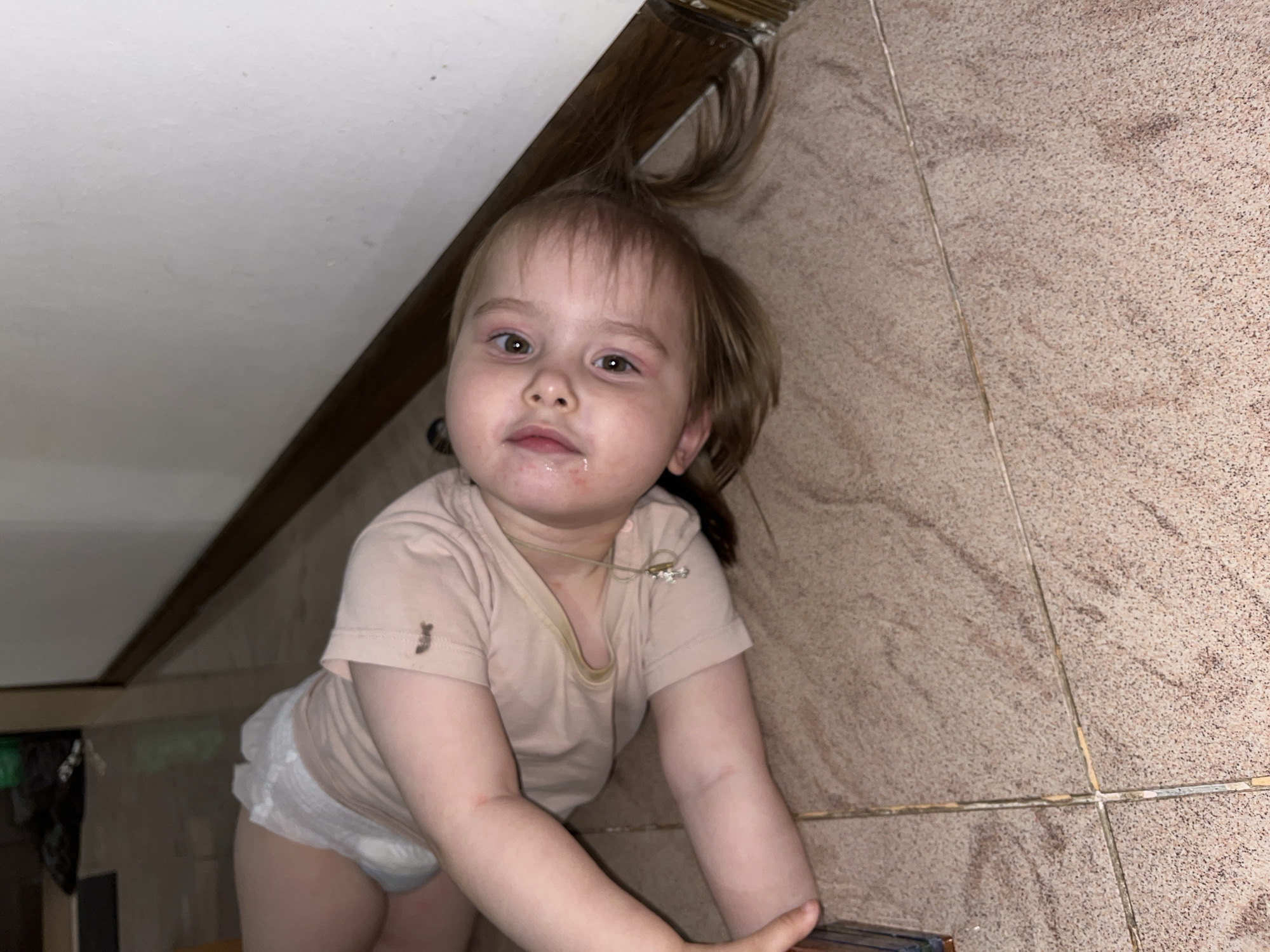 A young child, likely a toddler, is pictured from a low angle. The child has light brown hair styled in a messy bun that stands up from their head, with some strands falling around their face. They have wide, brown eyes and a slightly open mouth with what appears to be some food residue around their lips. Their face shows some redness around the eyes and cheeks, and there are a few small red marks on their arms.
The child is wearing a light peach-colored t-shirt and a diaper. There are some smudges of dirt on the t-shirt, one on the left chest and another on the left sleeve. A thin, light-colored string is visible around the child's neck, possibly a necklace.
The setting appears to be indoors, with a textured, tiled floor that has a mottled brown and beige pattern. A portion of a wooden beam or edge is visible in the upper left and right corners of the image, suggesting a corner of a room or possibly a slanted ceiling. The lighting is dim, casting shadows and creating a somewhat moody atmosphere. The overall impression is that of a candid, everyday moment. No text is visible in the image. The time of day is not discernible, nor is the weather. The location context provided is Novokuybyshevsk, Russia.