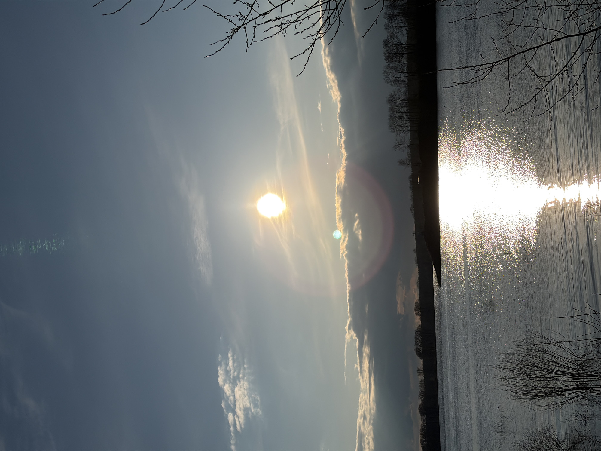 The image depicts a body of water, likely a river or a lake, reflecting sunlight. The sun is positioned high in the sky, partially obscured by thin clouds. The reflection of the sun on the water surface creates a sparkling effect. On the horizon, trees line the bank of the water. Bare tree branches are visible in the foreground, framing the scene. The overall lighting suggests either early morning or late afternoon. While it seems the image was taken in the city of Chaikovsky, Russia, no distinguishing landmarks provide explicit confirmation. The presence of trees, water, and sky suggests a natural environment, but further details are not discernible.