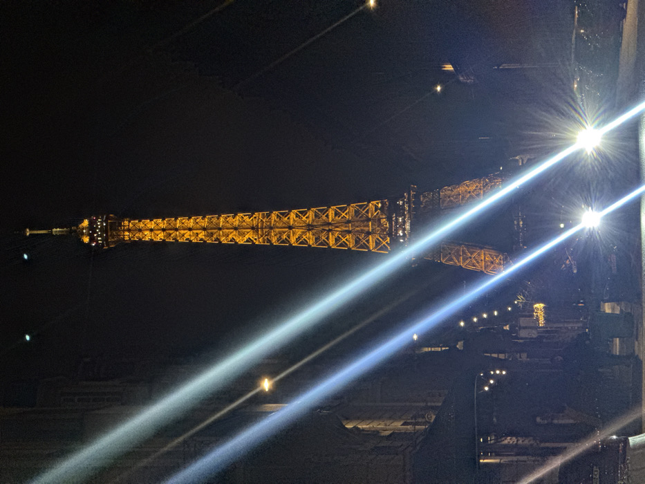 The image depicts a nighttime scene in Paris, France, dominated by the illuminated Eiffel Tower. The iconic lattice tower stands prominently in the center-right, glowing with a golden-yellow light against a dark, clear night sky. Its intricate structure is visible from its base to its spire, which is topped with a beacon.
In the foreground, a street or roadway is visible, characterized by intense lens flares originating from multiple vehicle headlights on the right and left sides. These flares manifest as bright, diagonal streaks of white and blue light traversing the lower half of the frame. Two distinct bright white light sources, presumably vehicle headlights, are centered in the lower-right foreground.
On the far left, a multi-story building with architectural details is dimly discernible. Below this building, an area with string lights and other faint illumination suggests an outdoor establishment, possibly a cafe or restaurant. Several indistinct human figures are present in the shadowed area of the street and near the building on the left, their forms largely obscured by darkness and light reflections, but appearing to be standing or walking. Parked or moving vehicles are present on the street, with a partial license plate ("TR 068" or similar) visible on a vehicle on the far left. Streetlights are also visible along the street, casting some ambient light. The overall environment is urban, dark, and characterized by artificial light sources.