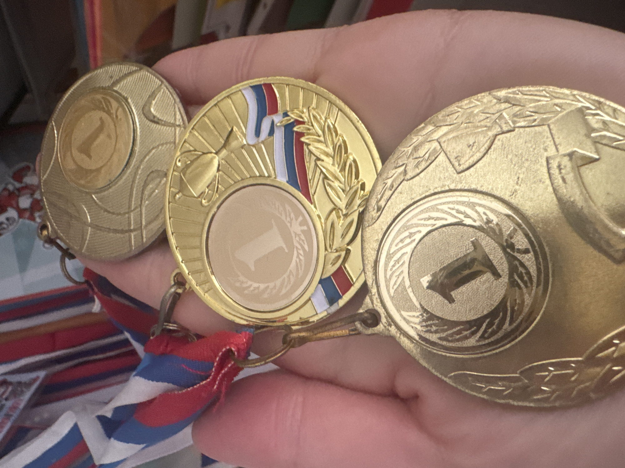 A hand holds three gold-colored medals, each bearing the numeral "1". The medals appear to be connected to a ribbon with blue, white, and red stripes. The setting of the photograph is not discernible from the image alone. There is no evidence that the medals are located in Votkinsk, Russia.