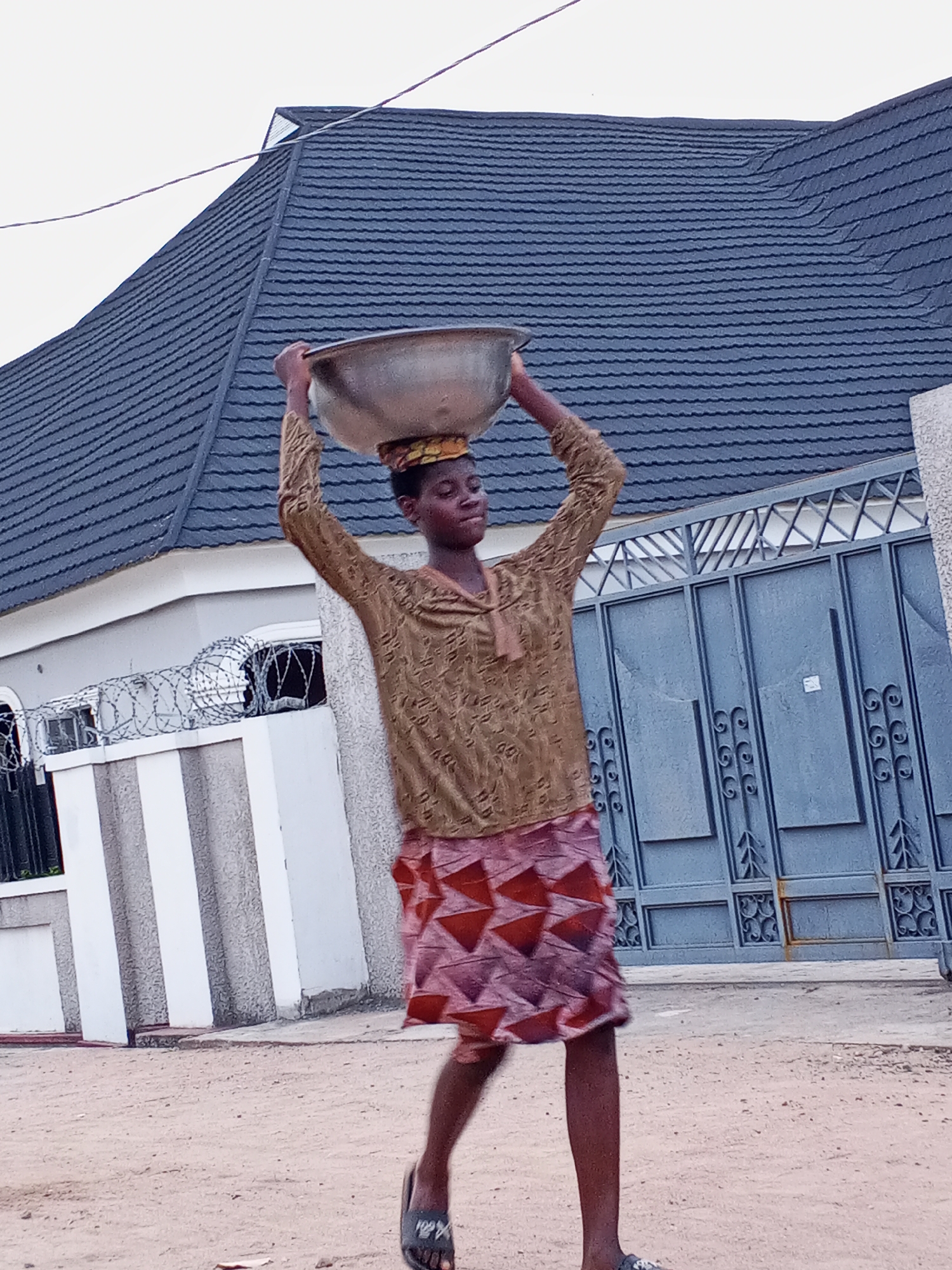 In Adonkolo, Nigeria, a woman walks on a sandy street carrying a large, round metal bowl on her head. She wears a brown patterned shirt, a patterned red and pink skirt, and sandals. Behind her is a residential building with a dark gray roof and a blue metal gate with ornate designs. The building is partially surrounded by a concrete wall topped with barbed wire. The sky is overcast.