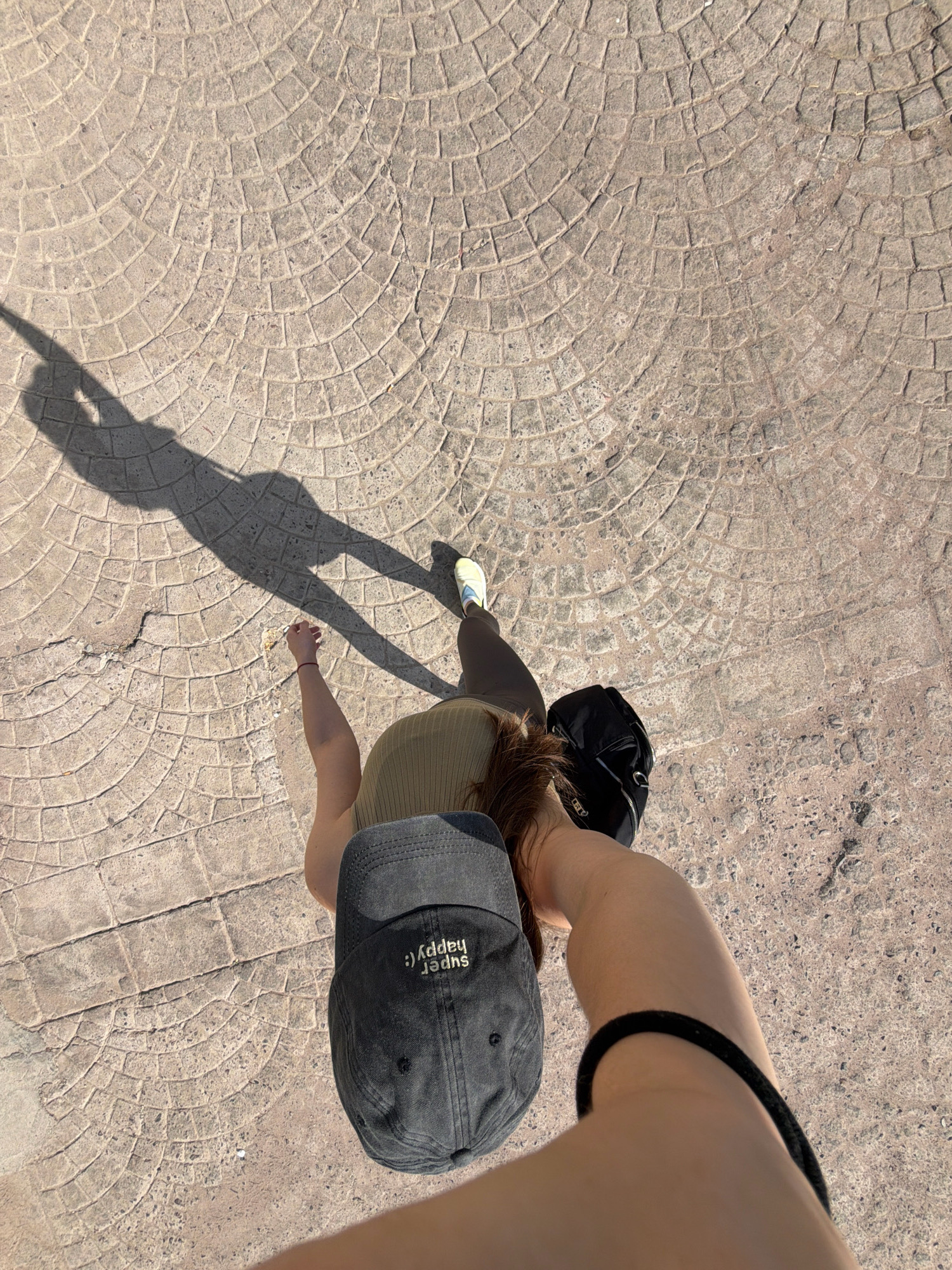 A first-person perspective shot from a slight angle shows a person standing on a circular patterned stone pathway in Sharm El Sheikh, Egypt. The person is wearing a green ribbed tank top, dark leggings, and light yellow athletic shoes. They are holding a black backpack in their right hand and a dark gray baseball cap with the words "super happy :)" embroidered on it is on their head. A long shadow of the person is cast on the pathway to their left. The pathway is made of individual square stones arranged in concentric circles. The lighting suggests it is daytime with the sun casting a strong shadow. The environment appears to be an outdoor urban setting.