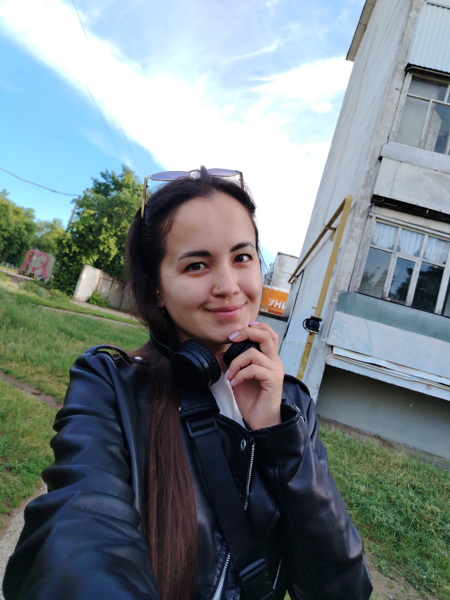 A young woman is featured in a selfie taken outdoors in Noviy, Russia, likely in a suburban or urban-fringe area. She has long, dark hair and is wearing a black leather jacket over a white top, with black over-ear headphones around her neck. Sunglasses are perched on her head. She has a subtle smile, a small jewel nose piercing, and small gold earrings. Her right hand is casually placed near her chin, where her light pink fingernails are visible.
The scene is set on what appears to be a grassy path, with green grass and trees visible to her left. In the distance on this side, there's a structure with a faint red, heart-like graphic or graffiti. To her right, a light-colored multi-story apartment building stands, featuring windows with white curtains and a prominent yellow gas pipe running vertically on its exterior. The sky above is clear and bright blue with wispy white clouds, indicating a sunny day, possibly late afternoon or early evening due to the soft light.
The woman appears relaxed and cheerful as she takes the self-portrait. In the background, behind her left shoulder, a partially obscured orange sign displays the Cyrillic letters "УНИ".