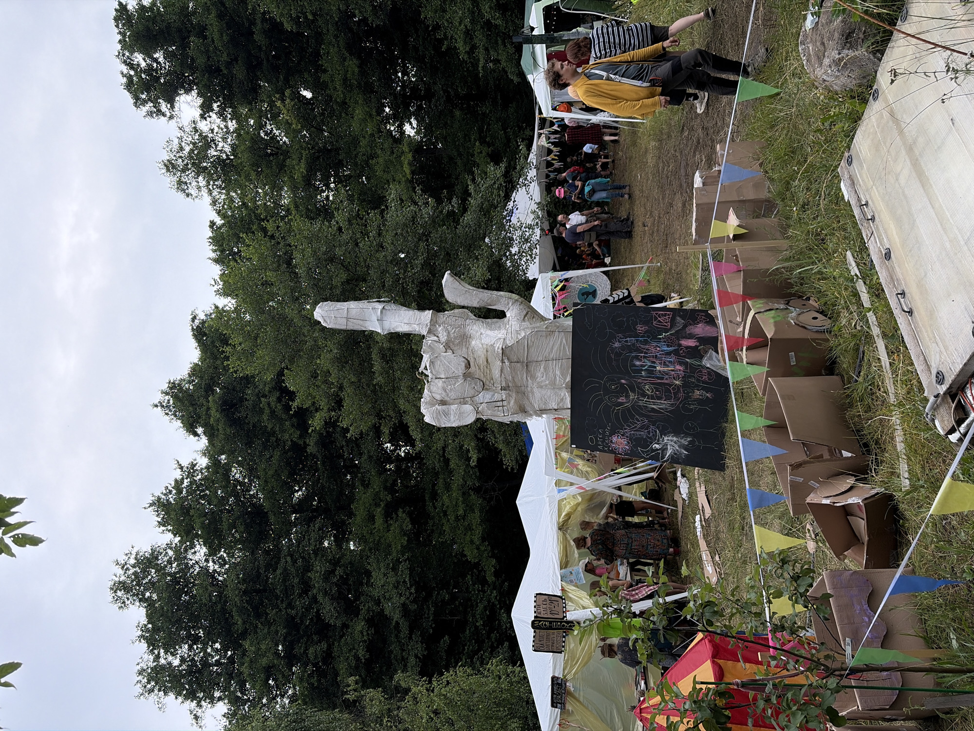The image depicts an outdoor gathering, possibly a festival or market, in a grassy area surrounded by trees. In the foreground, there are several cardboard boxes, some of which are open and contain items. A string of colorful pennant flags is stretched across the scene. To the left, a small, tent-like structure with red and yellow stripes is visible.

Dominating the center of the image is a large, abstract sculpture resembling a hand giving the middle finger, constructed from white material, possibly paper or fabric, wrapped around a frame. A blackboard with chalk drawings is placed in front of the sculpture.

In the background, several white tents are set up, with people milling about and interacting. Some individuals are standing, while others are seated. The environment appears to be a park or a wooded area on an overcast day. The text on the blackboard and signs on the tents are not legible in this resolution. The overall scene suggests a casual, communal event.

There is no information in the image or provided context to confirm the location as Il'inskiy Pogost, Russia.