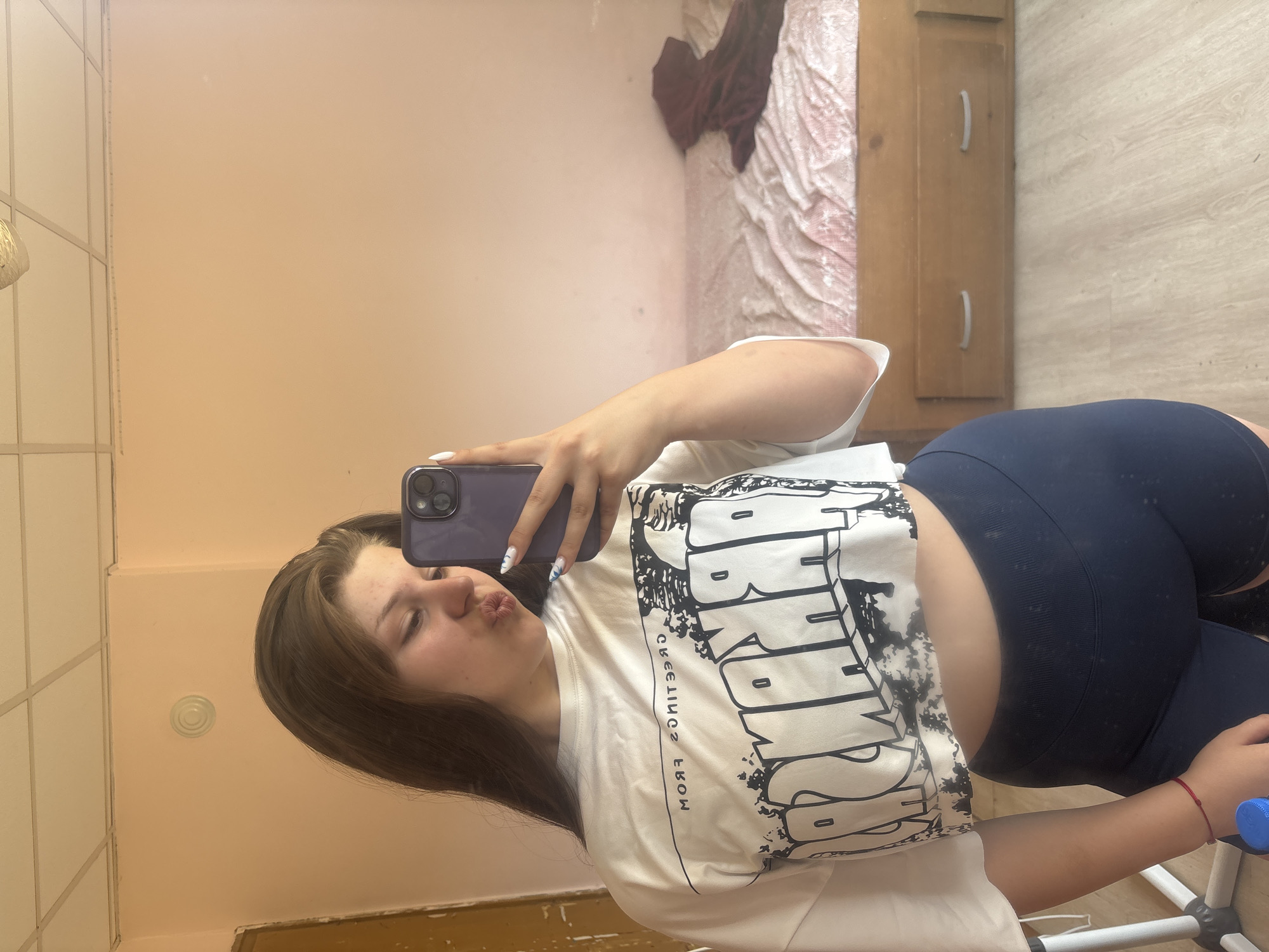 A young woman with long brown hair is taking a mirror selfie in a room with pale pink walls. She is wearing a white cropped t-shirt with a black graphic print and dark blue athletic shorts. Her left hand is holding a purple smartphone, and her right hand is visible. She is making a kissing face. In the background, there is a bed with a pink and white patterned comforter and a wooden headboard with drawers. To the left of the frame, part of a white object, possibly a tripod, is visible. The floor is light-colored wood. This image is not from Prodanovtsi, Bulgaria, as there are no specific visual cues in the image to suggest this location.