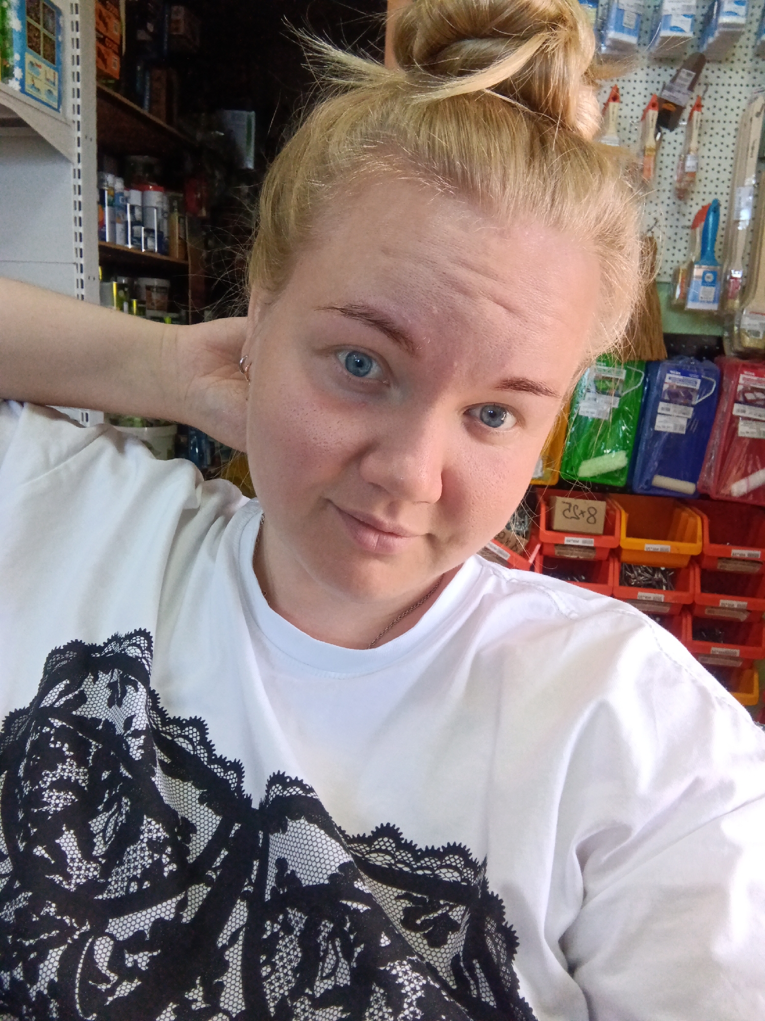 A young woman with blonde hair pulled up in a bun and blue eyes is taking a selfie. She is wearing a white t-shirt with a black lace pattern on the front. Her left arm is bent and her hand is behind her head. The background appears to be a retail store, possibly a hardware or craft store, with shelves stocked with various items, including boxes and tools. The environment is indoors and appears to be well-lit. There is no information to determine the specific location in the city of Фоки, Russia.