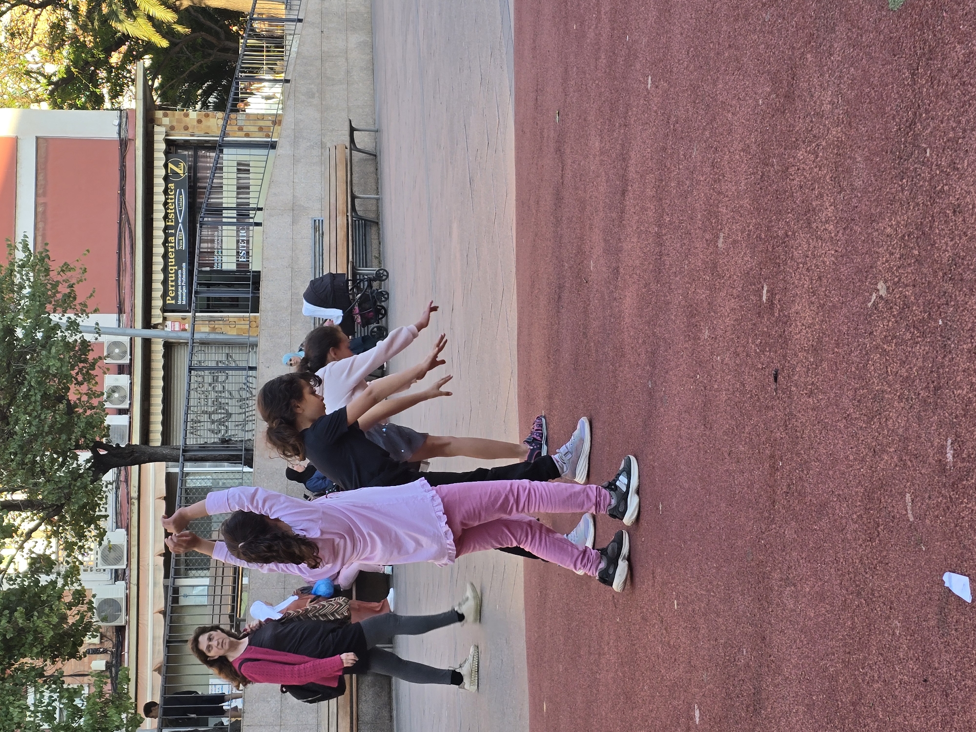 The image depicts a group of four females in Cornellà De Llobregat, Spain, engaged in what appears to be a stretching or yoga exercise in a public square. They are lined up on a dark red surface, possibly a running track or designated area for recreation. The first person in the line is an adult, likely a teacher or instructor, dressed in a black top and a pink sweater. The other three are girls who are stretching their arms above their heads. Behind them, there is a sidewalk with benches, trees, and a building with awnings and signs. The sign displays "Perruqueria I Estètica" and another vertically written word "Estetic". Several air conditioning units are fixed on the building's exterior wall. The square looks urban with a mix of trees and infrastructure. A stroller and a few other people are visible in the background.