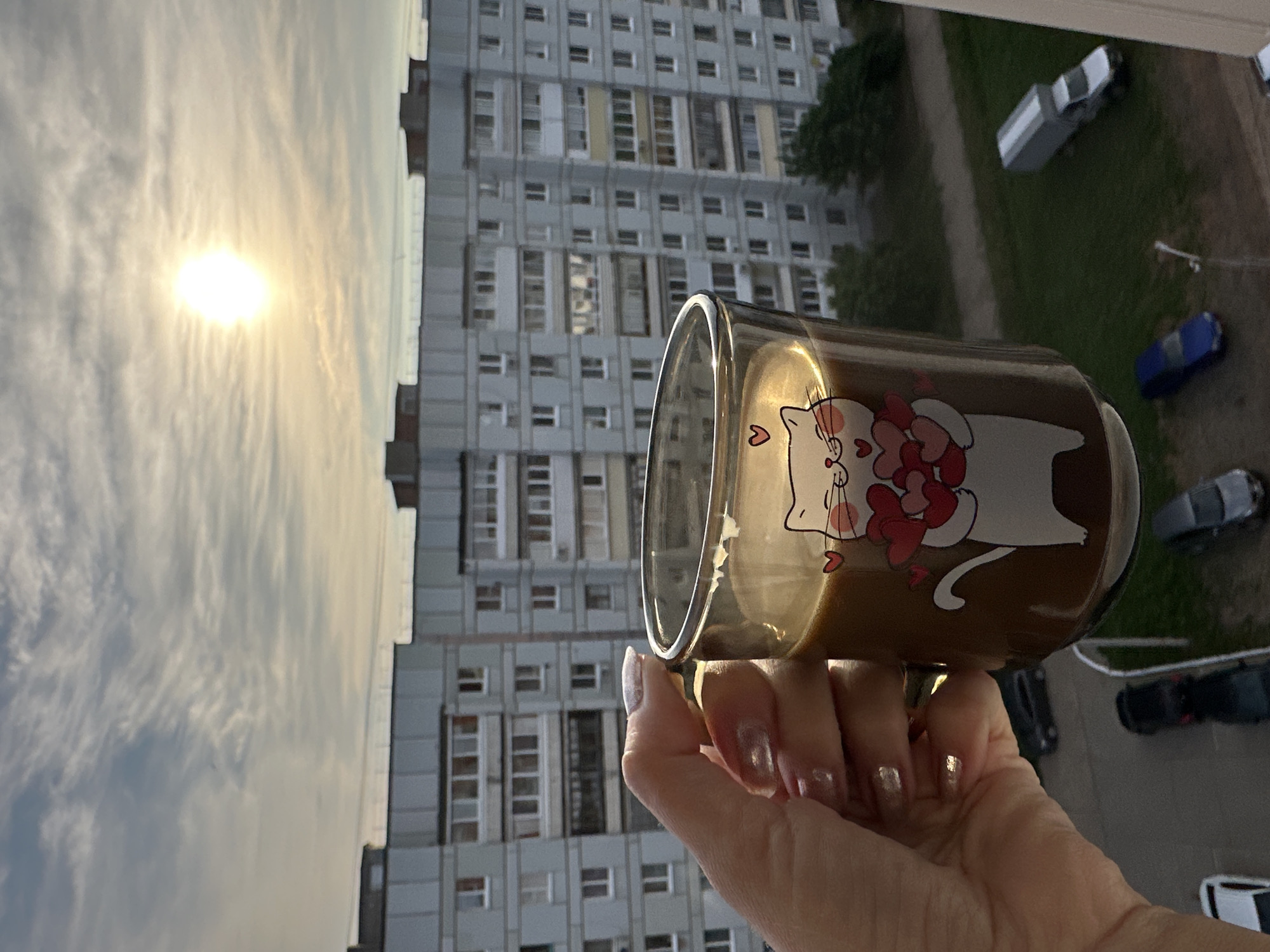The image, originally rotated 90 degrees counter-clockwise, captures a personal moment against an urban backdrop. A hand with manicured, light-colored nails holds a clear glass mug containing a brown liquid, likely coffee or tea. The mug is decorated with a white cartoon cat, featuring rosy cheeks and holding several red hearts, with an additional small heart symbol near its head.
The scene unfolds from what appears to be a high vantage point, possibly an apartment window. In the midground, a large, multi-story residential building with a repetitive pattern of windows and light-colored facades is prominent, characteristic of post-Soviet architecture, aligning with the provided context of Chaikovsky, Russia.
The background is dominated by a bright sun shining through a thin layer of clouds, suggesting either early morning or late afternoon. Below the building, ground level reveals a grassy area, a dirt path, and several parked cars, including a white and grey van, a blue car, and a black vehicle. Green foliage from trees or bushes is also visible. The image conveys a sense of peaceful contemplation, contrasting the intimate detail of the mug with the expansive view of the city and sky.