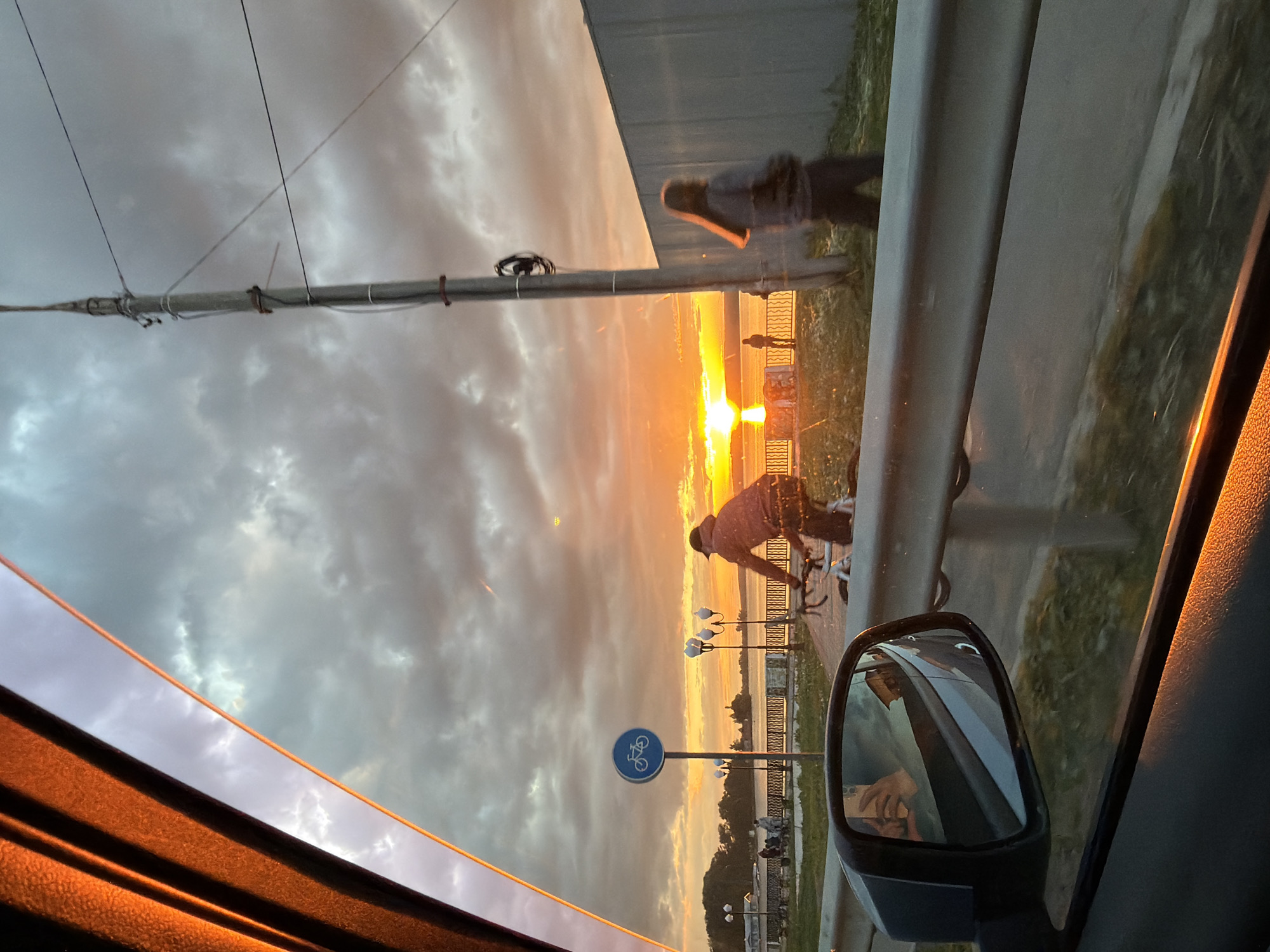 Captured from inside a vehicle, the photograph presents a view of Votkinsk, Russia at sunset. The sky is filled with dramatic clouds, illuminated by the setting sun. In the foreground, a bicyclist rides along a paved path adjacent to a river embankment. A bicycle lane sign is visible. Further along the embankment, people are gathered, seemingly watching the sunset. The scene is partially framed by elements of the car's interior, including the window frame and side-view mirror, in which a reflection of the car’s occupant is visible. A barrier stands along the path. Power lines are visible in the upper left of the image.