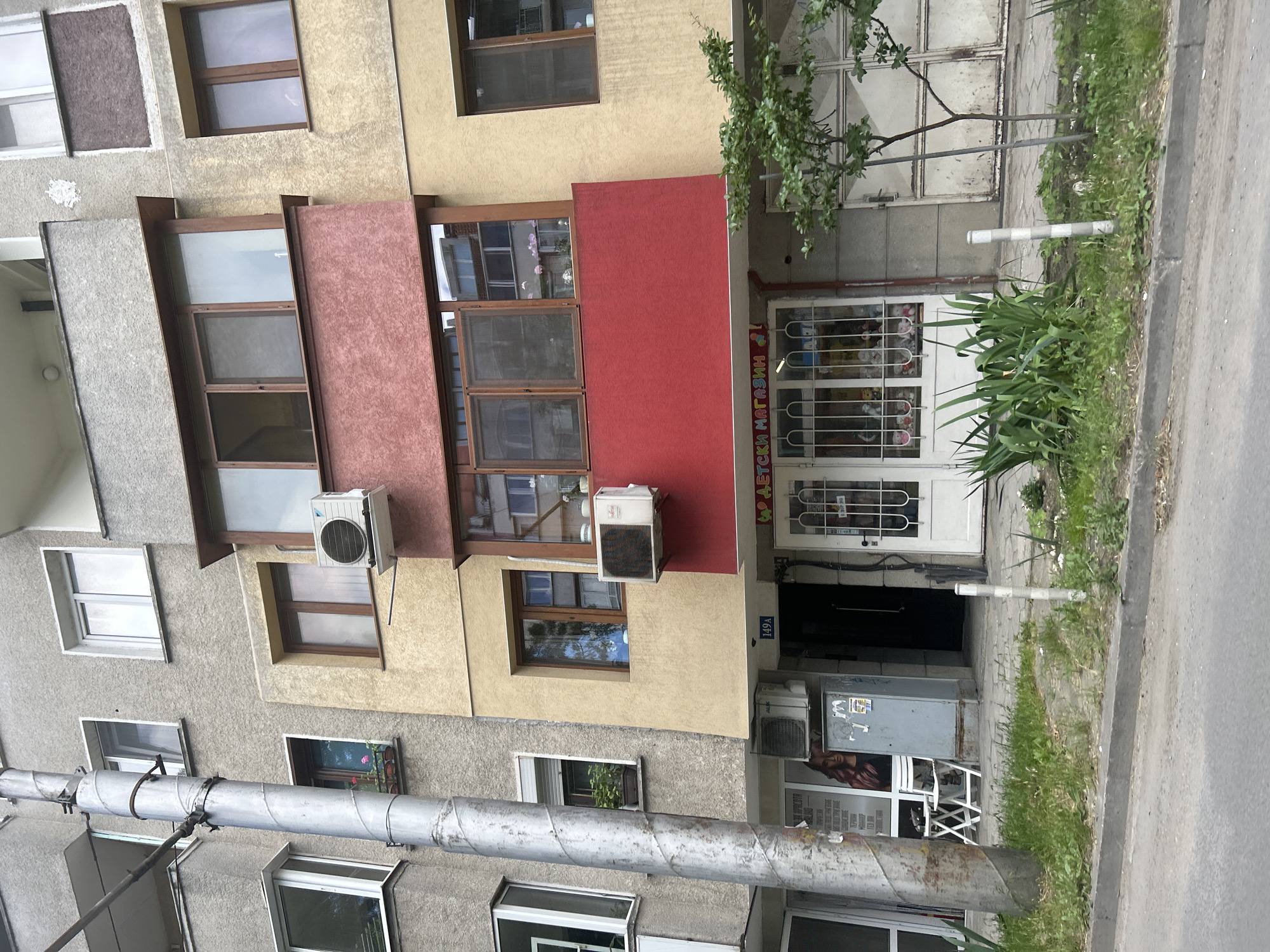 The image depicts the facade of a multi-story apartment building with a storefront at ground level, located on a street in Varna, Bulgaria. The building has a combination of gray, beige, and yellow exterior walls, with several windows and balconies. The storefront features a large glass window displaying toys and other merchandise, with a red awning above that reads "ДЕТСКИ МАГАЗИН" (Children's Store). A white door is visible to the right of the storefront. In front of the building, there is a sidewalk with some greenery and a tree. A utility pole stands prominently on the left side of the image. The street in the foreground appears to be paved with asphalt.