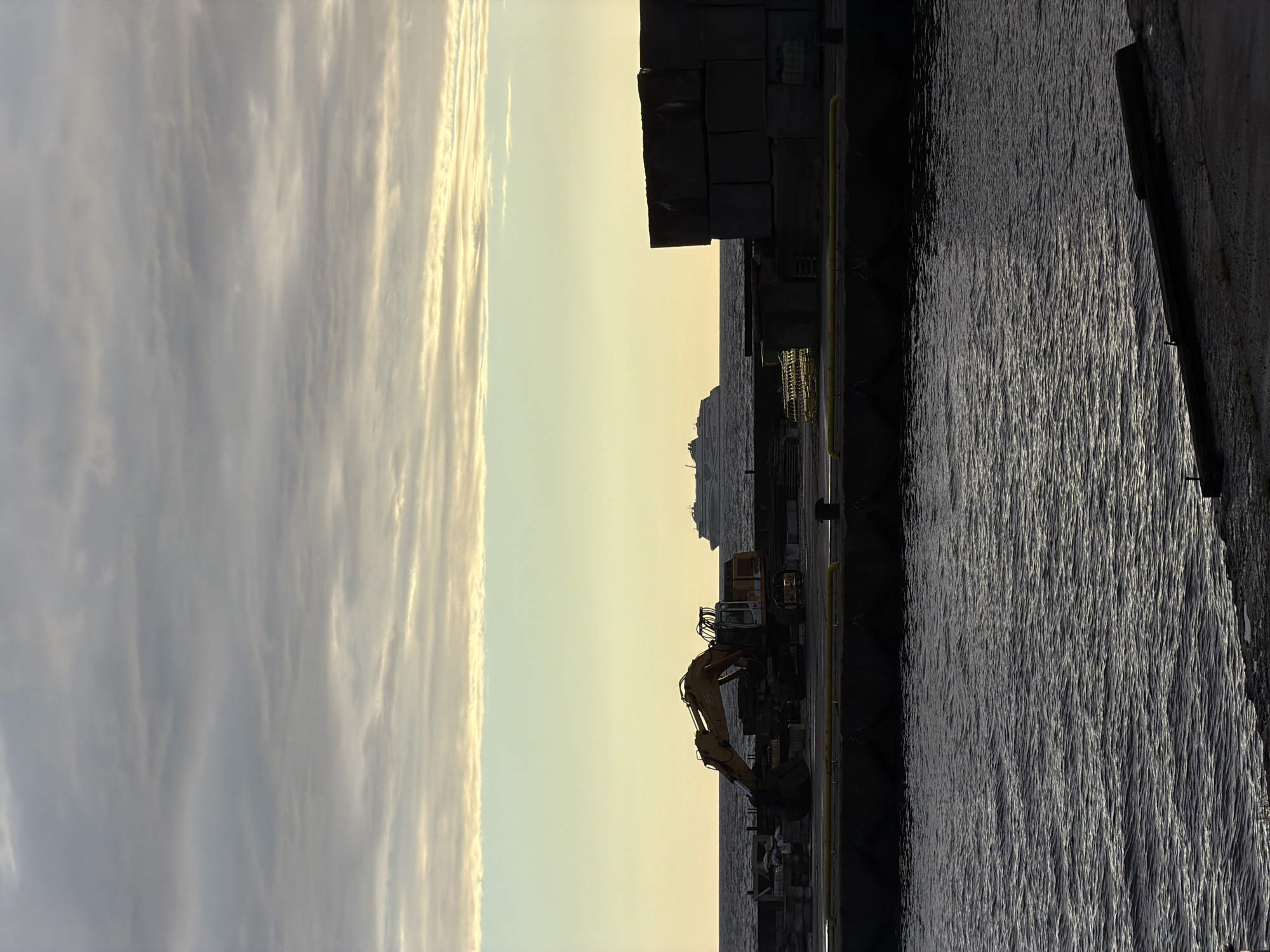 This media file captures a harbor scene at dawn in Viimsi, Estonia. The image is oriented vertically, with a wide expanse of clouds dominating the left side, appearing wispy and illuminated by the rising sun. To the right, the sky transitions to a pale yellow and then a soft orange, characteristic of early morning light.
In the middle ground, a port or industrial area is visible. Large, dark, rectangular blocks are stacked prominently, possibly forming part of a structure or cargo. Below these blocks, a yellow barrier or railing runs horizontally. An excavator, predominantly yellow with a black cabin, is positioned on the dock. Its mechanical arm is extended downwards towards the water.
The right side of the image is filled with dark, rippling water, reflecting the subdued light from the sky. The surface of the water is textured with small waves, creating a shimmering effect. A distant ship or ferry is faintly visible on the horizon, adding to the maritime context. The overall atmosphere is calm and serene, indicative of the quiet start to the day. The lighting suggests it is either sunrise or sunset, with the colors leaning towards sunrise. There is no visible text in the image.