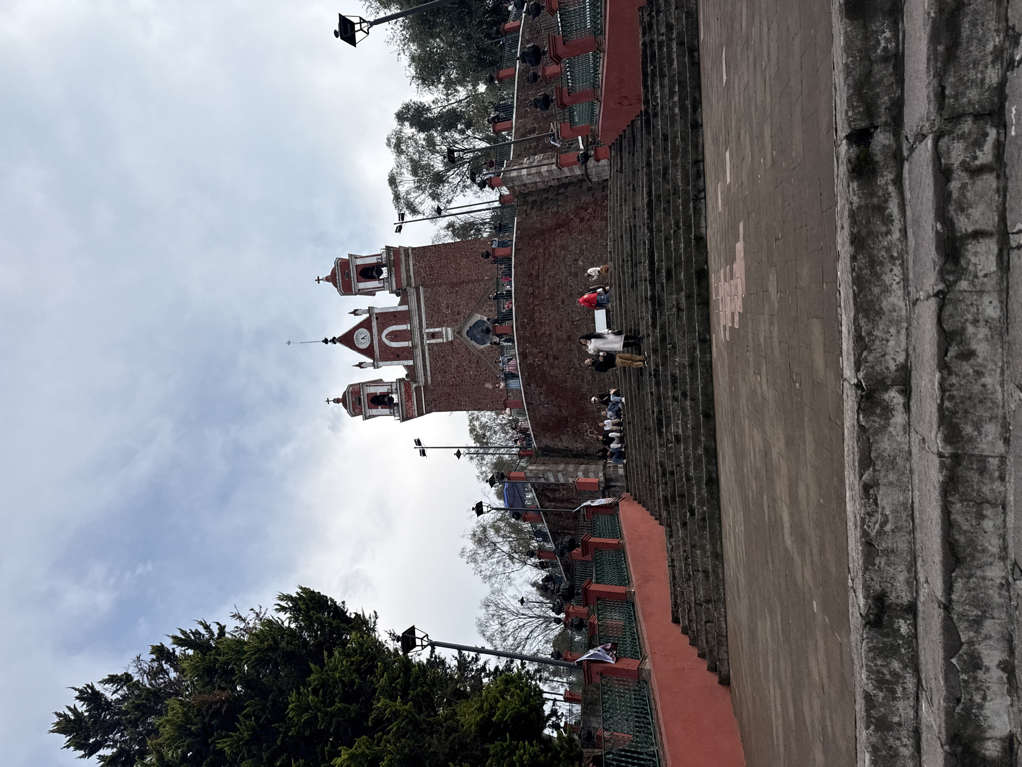 The image captures a view of the Ex Convento de San Miguel Arcángel, located atop the Cerro de Muguette in Metepec, Mexico. The red brick church with two bell towers and a clock tower dominates the frame. People are present on the steps leading up to the church. The stairs are made of stone. A red and green railing runs alongside the stairs. Street lamps are also seen along the steps. Trees are visible in the foreground. The sky is overcast.