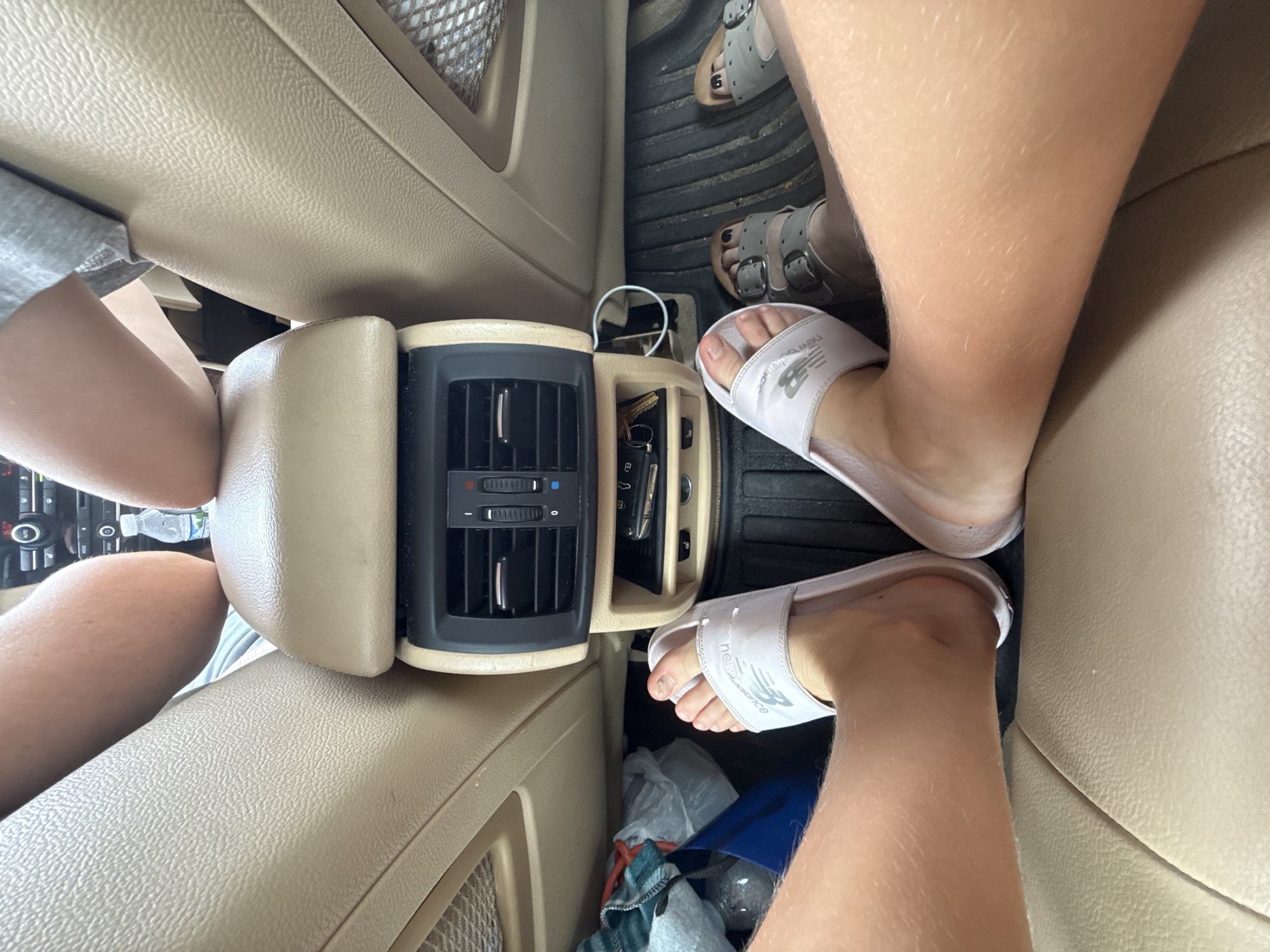 This is a close-up, low-angle, interior shot of a car, likely taken in Skokie, United States of America. The perspective is from the back seat, looking forward. Two pairs of feet are visible, one wearing light pink slides with a "New Balance" logo, and another wearing grey strap sandals. The floor mat is black and ribbed. In the background, there is a car's center console with air vents and some keys. To the left, a water bottle and some bags are visible. The overall impression is one of casual travel.