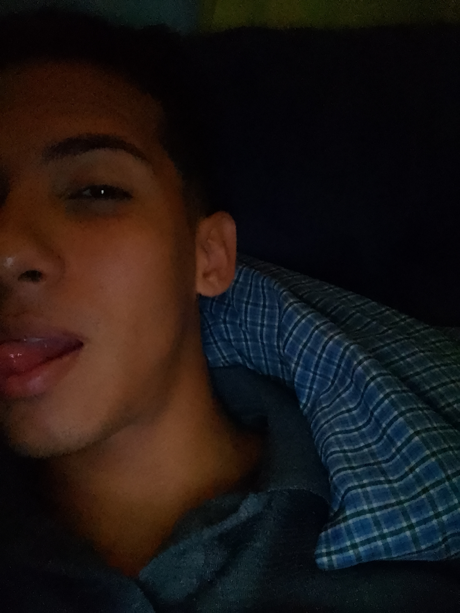 A person with dark skin and short dark hair is lying down, possibly in bed, with a blue and white plaid blanket around their shoulder. Their tongue is slightly sticking out. The background is dark, suggesting a low-light environment. No information about Pampatar, Venezuela is discernible from the image.