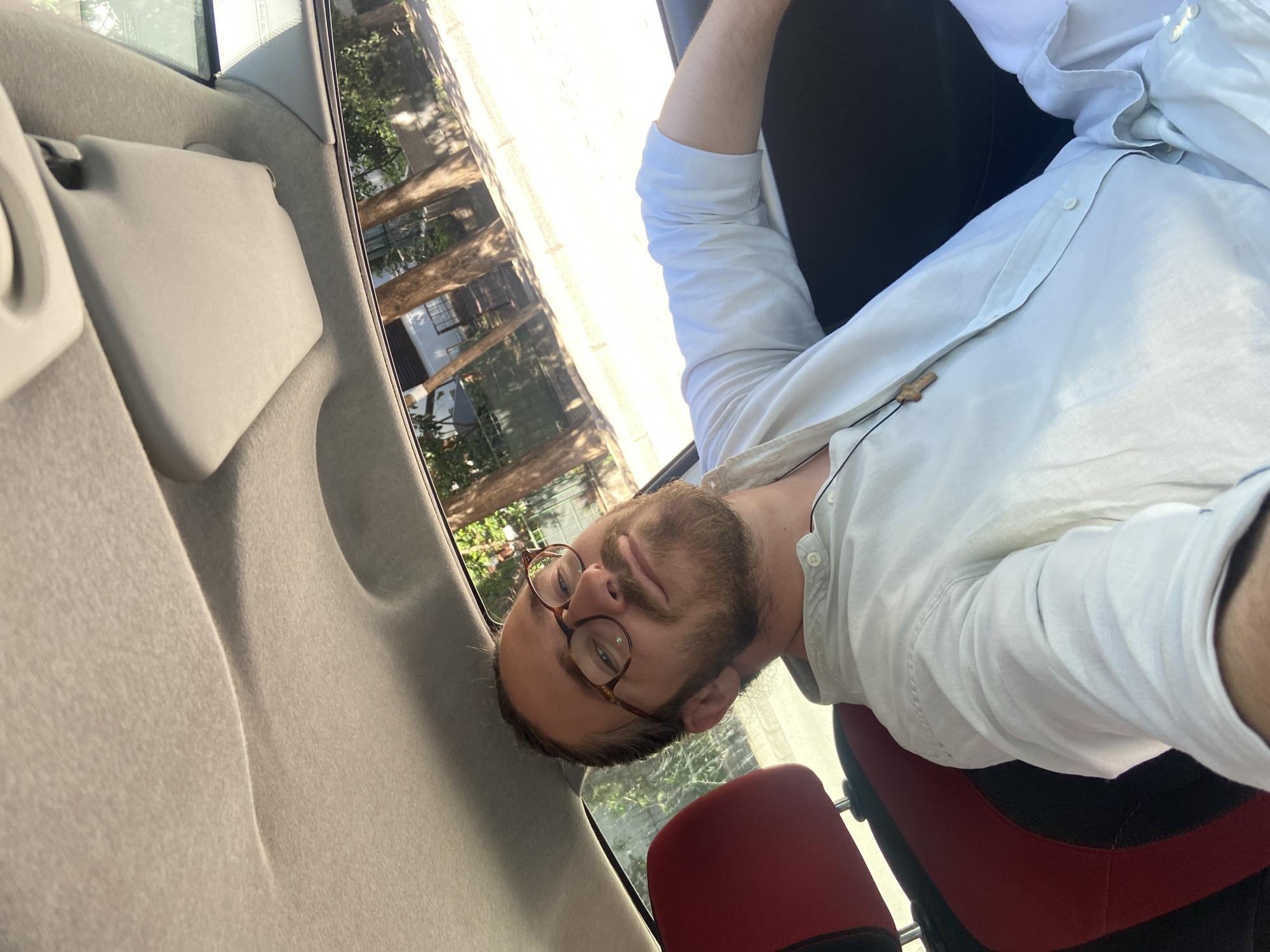 A man with a beard and glasses sits in the passenger seat of a car. He is wearing a white button-down shirt and black pants. The interior of the car is visible, including the gray upholstery of the car door and the red and black seat. The exterior of the car shows trees and buildings that match the architecture of Budva, Montenegro.