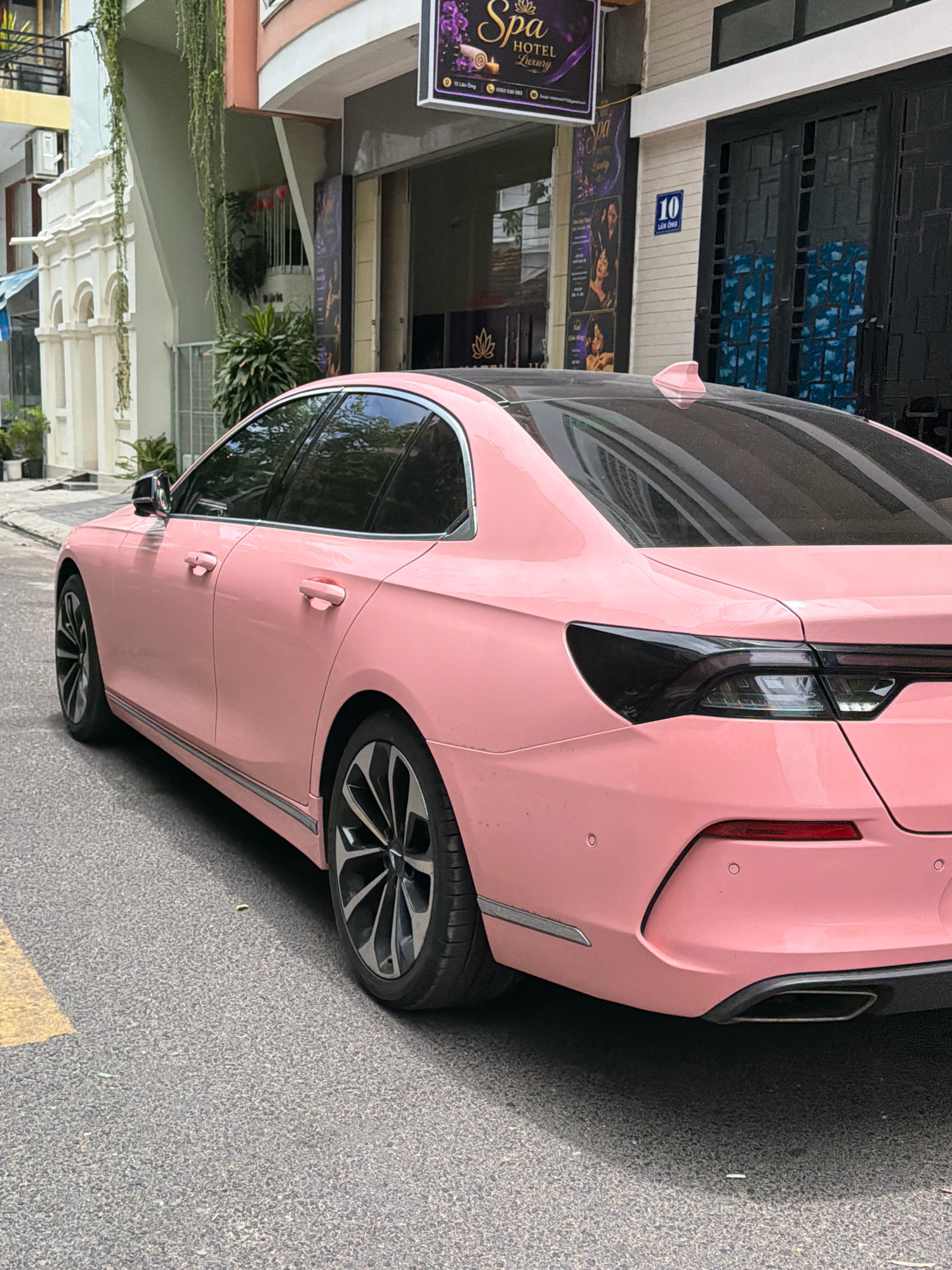 A pink sedan is parked on a street in Nha Trang, Vietnam. The car is viewed from the rear passenger side, showing its sleek design and modern taillights. The background features a building with a sign that reads "Spa Hotel Luxury." The street is paved with asphalt and has a yellow line painted on the side. The overall scene is typical of an urban environment in Vietnam.