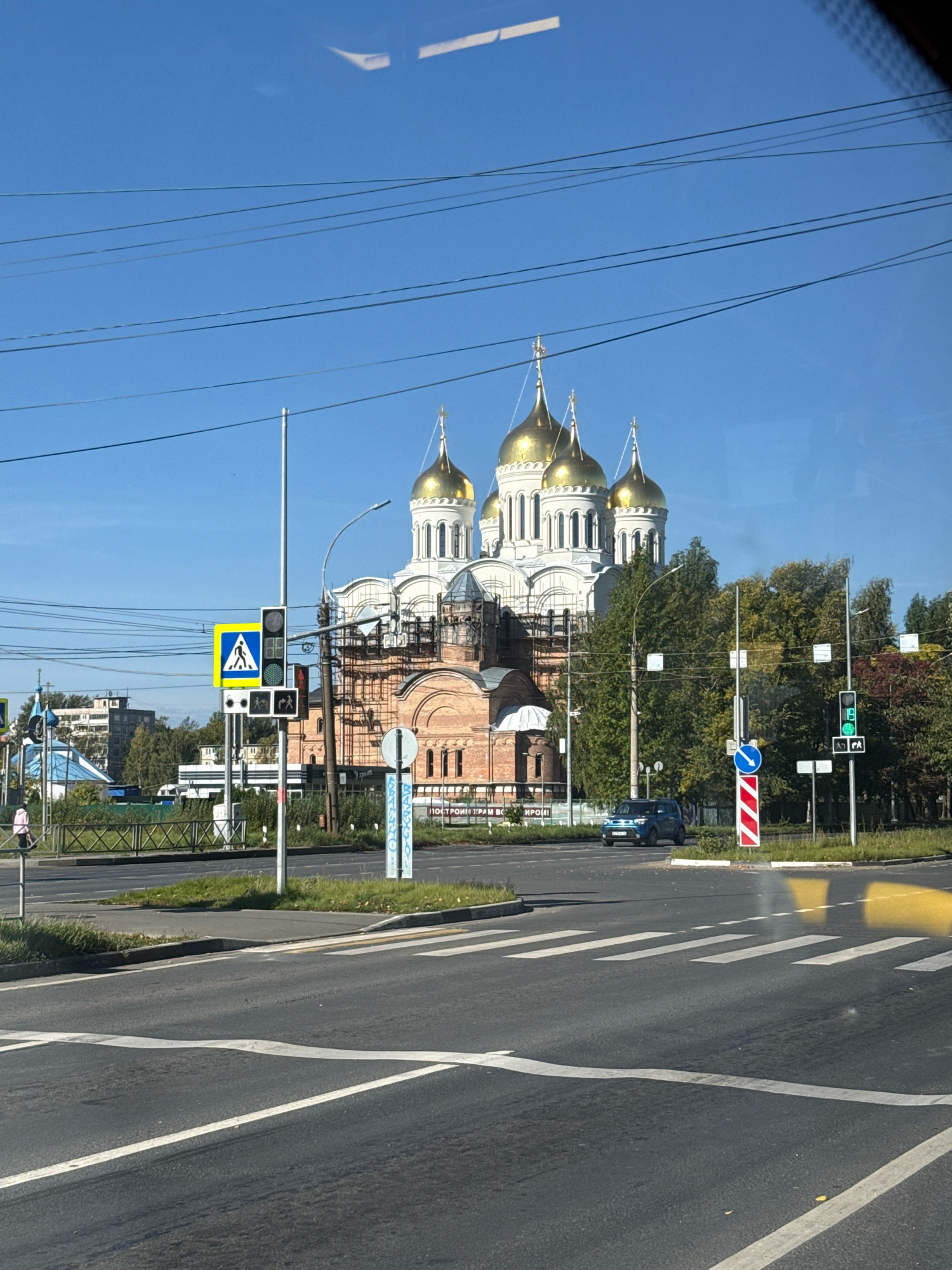 This image, taken from inside a vehicle, shows a wide asphalt road with multiple lanes and pedestrian crossings. A blue car is visible on the right side of the road. In the background, a large, ornate church with golden domes is under construction, with scaffolding visible. The sky is clear and blue. The scene appears to be in a city, with trees and buildings in the distance. This is the Church of the Resurrection of Christ in Rusanovo, Russia.