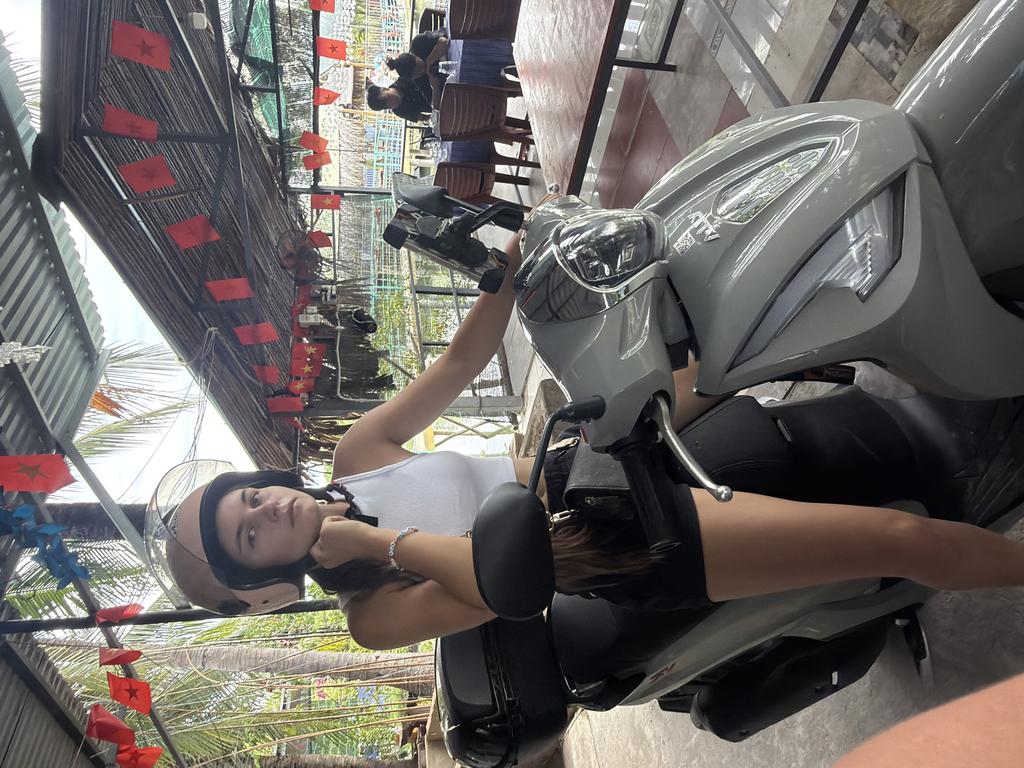 A young woman wearing a tan helmet, a white crop top, and black shorts sits on a grey scooter. She is looking towards the camera with her hand resting on her chin. The scooter is parked under a thatched roof structure adorned with red Vietnamese flags. In the background, several people are seated at tables in an outdoor dining area. The environment appears to be a restaurant or cafe near a body of water, possibly in Nha Trang, Vietnam, given the cultural context of the flags.
