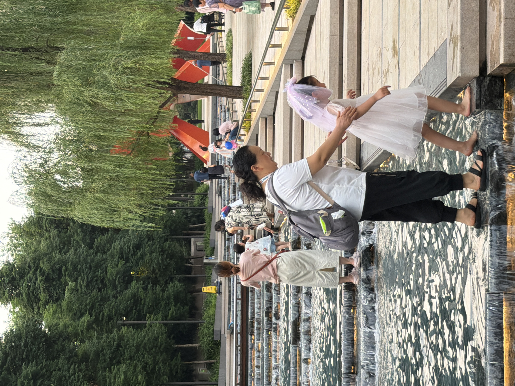 This image captures a public plaza in the Chongqing Grand Theatre Plaza, located in Yuzhong District, Chongqing, China. The scene features a multi-tiered stepped water fountain integrated into a paved urban landscape, surrounded by greenery and architectural elements.
In the foreground, a woman wearing a white short-sleeved top, black pants, and black sandals with a grey backpack, is assisting a young girl. The girl, dressed in a white short-sleeved dress with a sheer pink veil-like headpiece, is barefoot and stepping onto a wet, dark-colored step. The woman has her right hand gently placed on the girl's left arm/shoulder area, looking down at the girl.
To the left in the midground, another woman with her back to the camera, wearing a light pink t-shirt and a long light-colored skirt, stands in the shallow water of the fountain. Her hair is tied in a bun, and she carries a red shoulder bag. A small child in a patterned light-colored outfit is visible next to her, also in the water. Further back and to the right of this group, a woman in a patterned dress is seen bending down near another child by the water. The water feature consists of multiple wide, shallow steps over which water cascades, reflecting ambient light.
The background reveals a wide, light-colored paved plaza area with stairs leading upwards. A prominent weeping willow tree with long, hanging green branches is situated on the right side of the plaza. Behind this tree and across the plaza, large, abstract, bright orange geometric sculptures, characteristic of the "Cloud Gate" sculptures at Chongqing Grand Theatre Plaza, are visible. Various individuals are dispersed throughout the background, walking or standing on the plaza. On the far left, beyond the fountain, a line of dense green trees creates a backdrop, with streetlights visible among them. A yellow "wet floor" cone is visible far in the distance on the left side near the trees. The lighting suggests daytime, possibly late afternoon or early evening, with soft, diffused light.