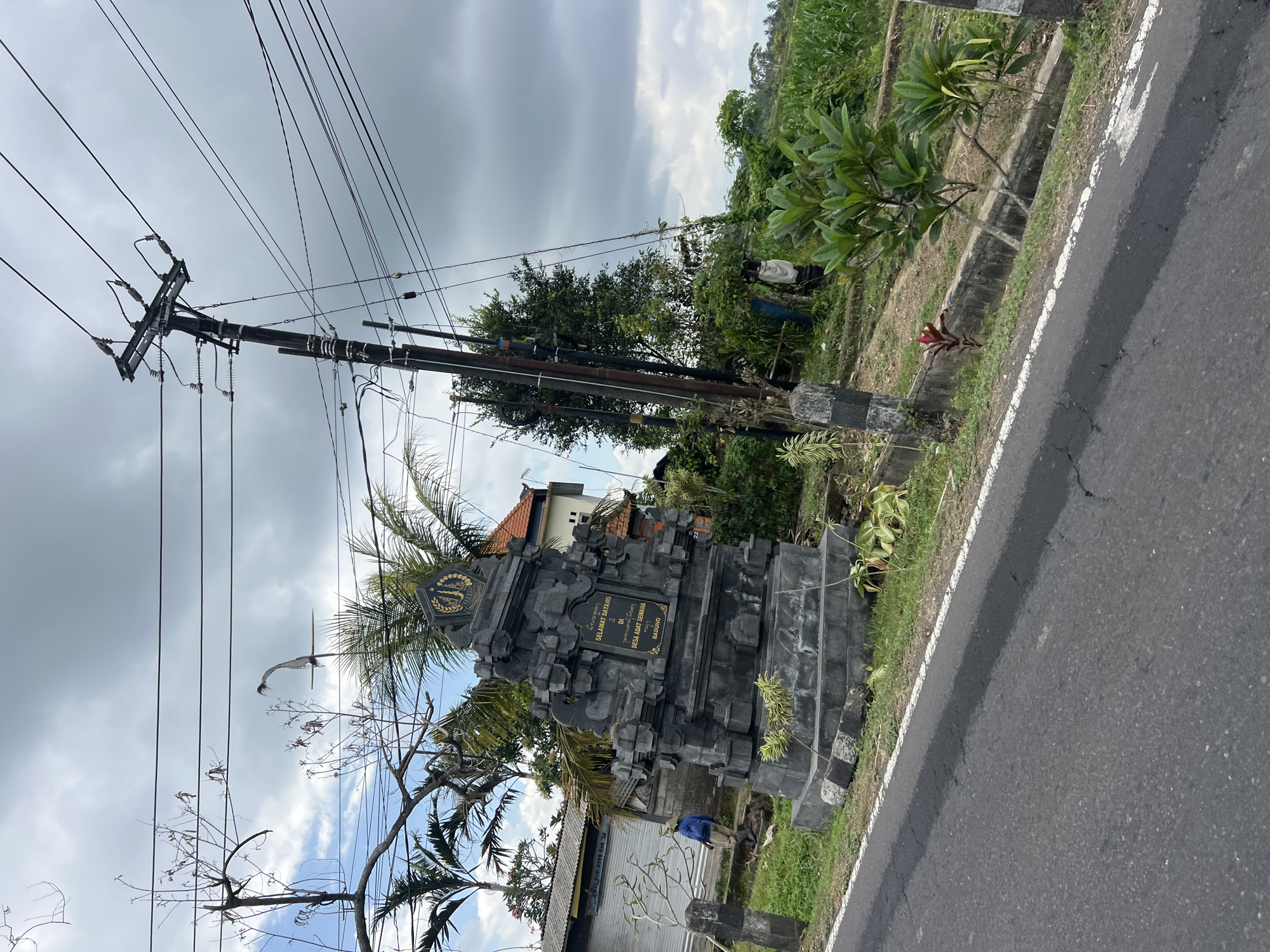 An outdoor, ground-level shot captures a stone monument on the side of a road, likely in Abiansemal, Indonesia, given the Balinese inscription. The monument, featuring intricate carvings, stands against a backdrop of lush greenery, palm trees, and a cloudy sky. A utility pole with multiple wires is prominent in the frame, positioned behind and to the right of the monument. The road in the foreground shows signs of wear with some cracks. A person dressed in a blue jacket is visible to the left of the monument, appearing to be walking away. In the background, a building with a tiled roof is partially visible. To the right of the monument, a field of crops, possibly corn, stretches into the distance.