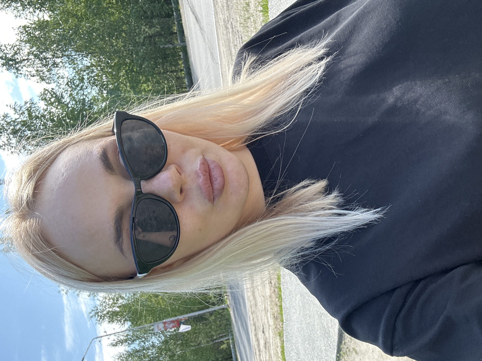 A blonde woman wearing black sunglasses and a black t-shirt is taking a selfie outdoors. She has light skin and full lips. Her hair is light blonde and falls around her shoulders. She is wearing black sunglasses that cover her eyes and the top part of her nose. The background shows a street or road with trees on the right side and a blue sky with some clouds overhead. The lighting suggests it is daytime and sunny.