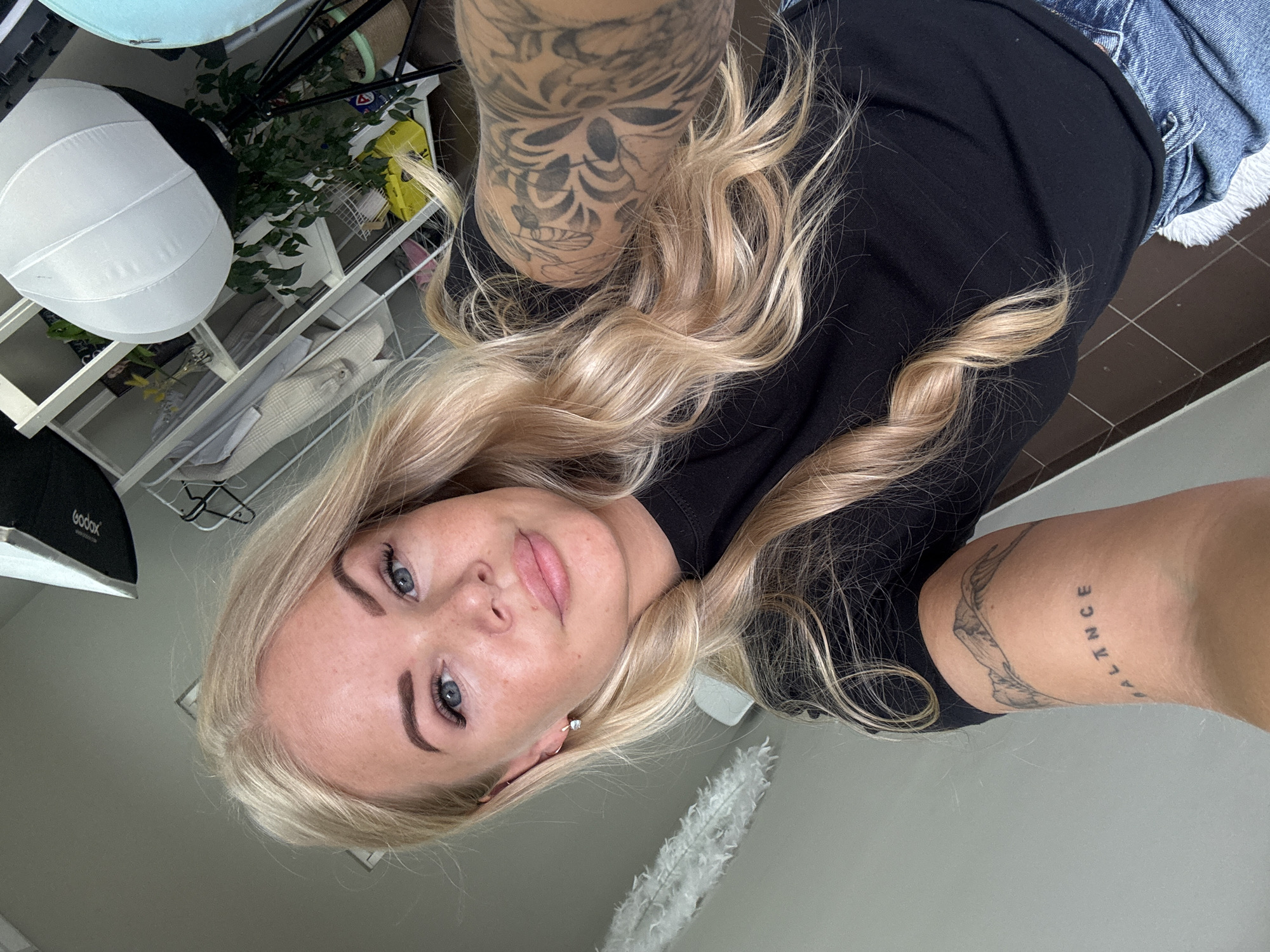 A selfie-style photograph captures a fair-skinned individual with blonde, wavy hair. She has blue eyes and is wearing a black shirt and denim jeans. Tattoos are visible on her arms. One arm has a tattoo with a mountain range design along with the word "BALANCE". There is a tattoo on the other arm with skull design. The background includes a shelving unit with various items, including what appears to be photography equipment and a plant. The location is unknown.