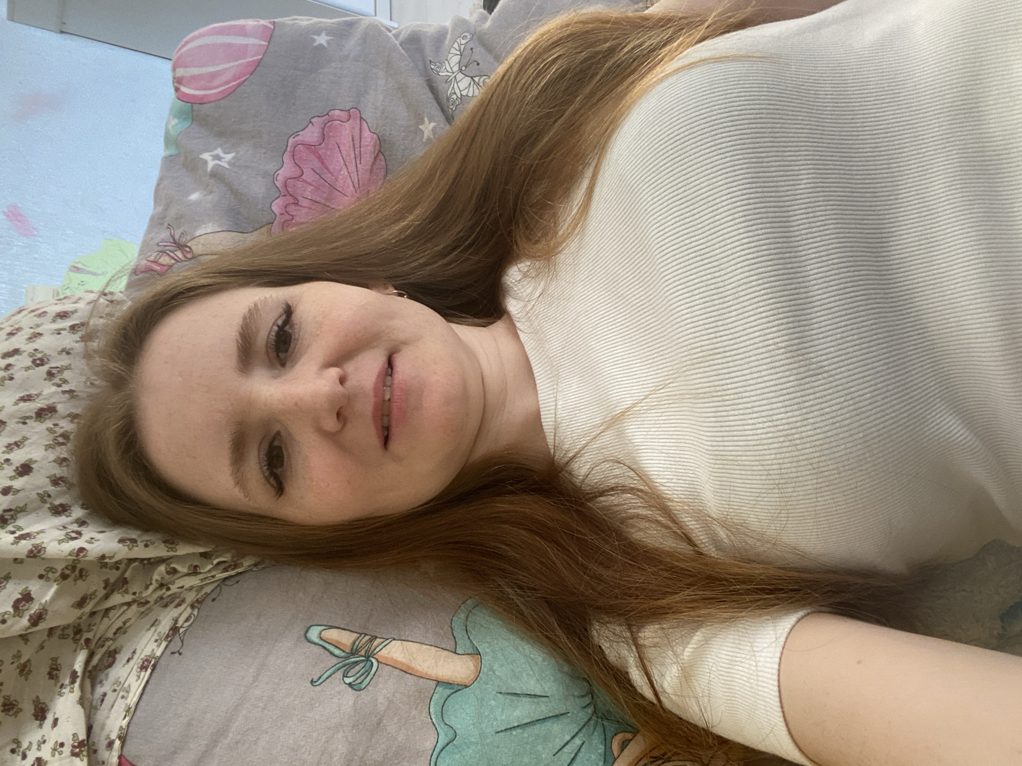 A young woman with long auburn hair is lying on patterned bedding. She is wearing a white ribbed top. The bedding includes a floral-patterned pillowcase and another with a design featuring ballet slippers, shells, and stars. The location is in Chaikovsky, Russia.