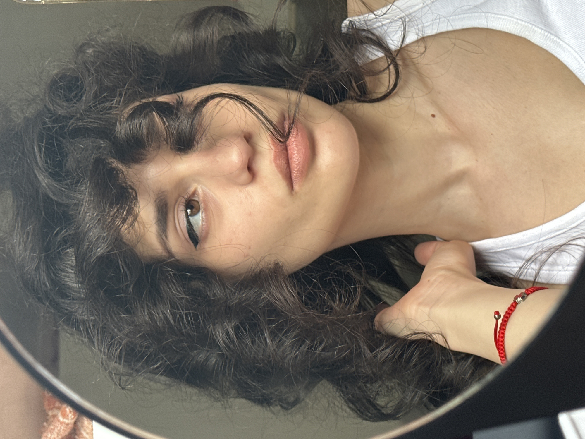 A woman with dark, curly hair is reflected in a round mirror. She is wearing a white tank top and a red bracelet on her left wrist. The reflection shows her face from the nose up, with her hair falling across her face. Her eyes are brown, and she is wearing eyeliner. Her lips are slightly parted. The background is blurred and appears to be indoors. There is no identifiable location in the city of Sofia, Bulgaria, mentioned or depicted in the image.