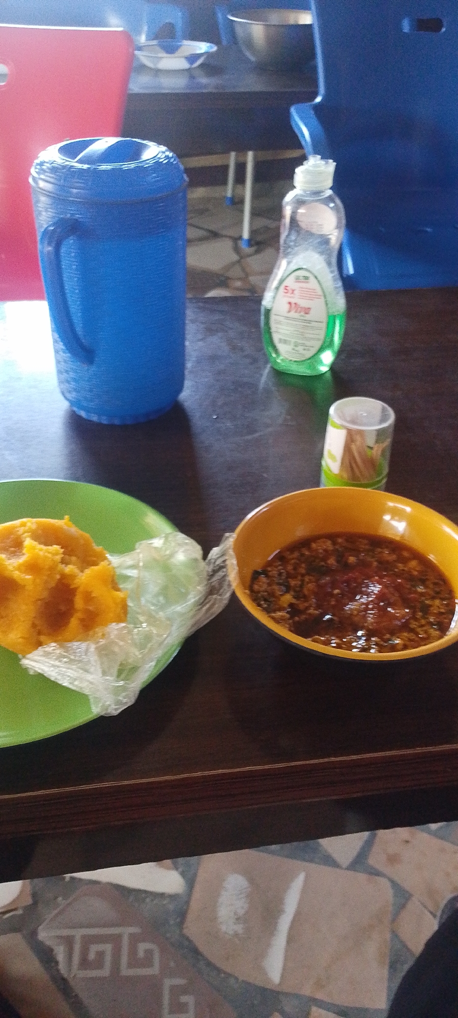 The image shows a table in Lokoja, Nigeria. On the table are a green plate with what appears to be yellow pounded yam partially covered in plastic wrap, a yellow bowl containing a stew-like sauce, a container of toothpicks, a blue water pitcher, and a bottle of green dish soap. A red plastic chair is partially visible behind the water pitcher, and a blue plastic chair is visible in the background. The table is dark brown. The floor beneath the table has a patterned tile design. A person's leg is visible in the lower right corner.