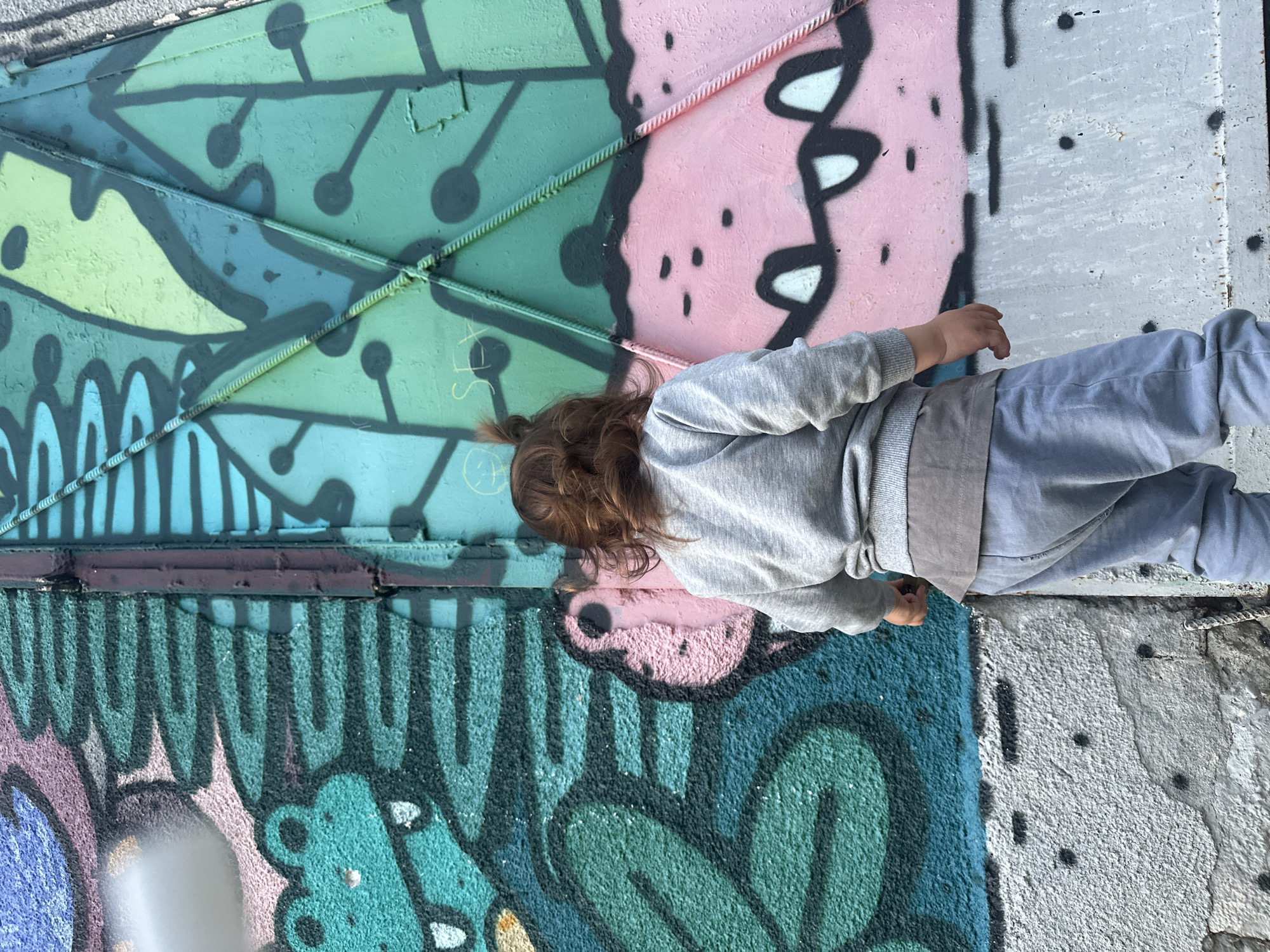 In Sofia, Bulgaria, a young child with curly brown hair stands facing a vibrant, graffiti-covered wall. The child, wearing a gray sweatshirt and light blue pants, is viewed from the back. The wall displays a colorful mural featuring cartoonish creatures and abstract designs in shades of green, pink, and blue. The mural extends across what appears to be a metal structure, possibly a gate or door, evidenced by the visible seams and textured surface. The lower portion of the wall is a rough, concrete surface with black spray-painted dots.
