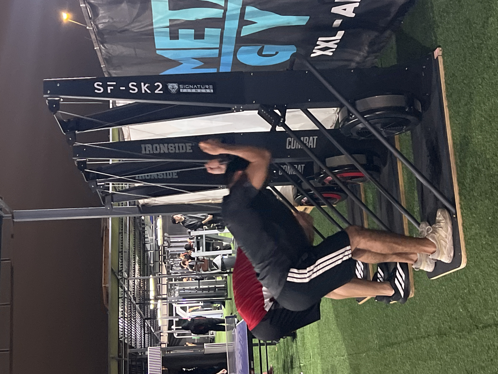 Two men are performing exercises on a gym floor with artificial turf, possibly at a gym in Arica, Chile. The man in the foreground is wearing a black t-shirt and black shorts with white stripes, and the man behind him is wearing a red t-shirt and black shorts. They are both using exercise machines that appear to be rowers or similar equipment. The machines have black frames and circular fans at the bottom. In the background, other gym equipment and people can be seen. A banner with the text "METAL GYM" is visible to the right of the frame. The lighting suggests it is evening or night.