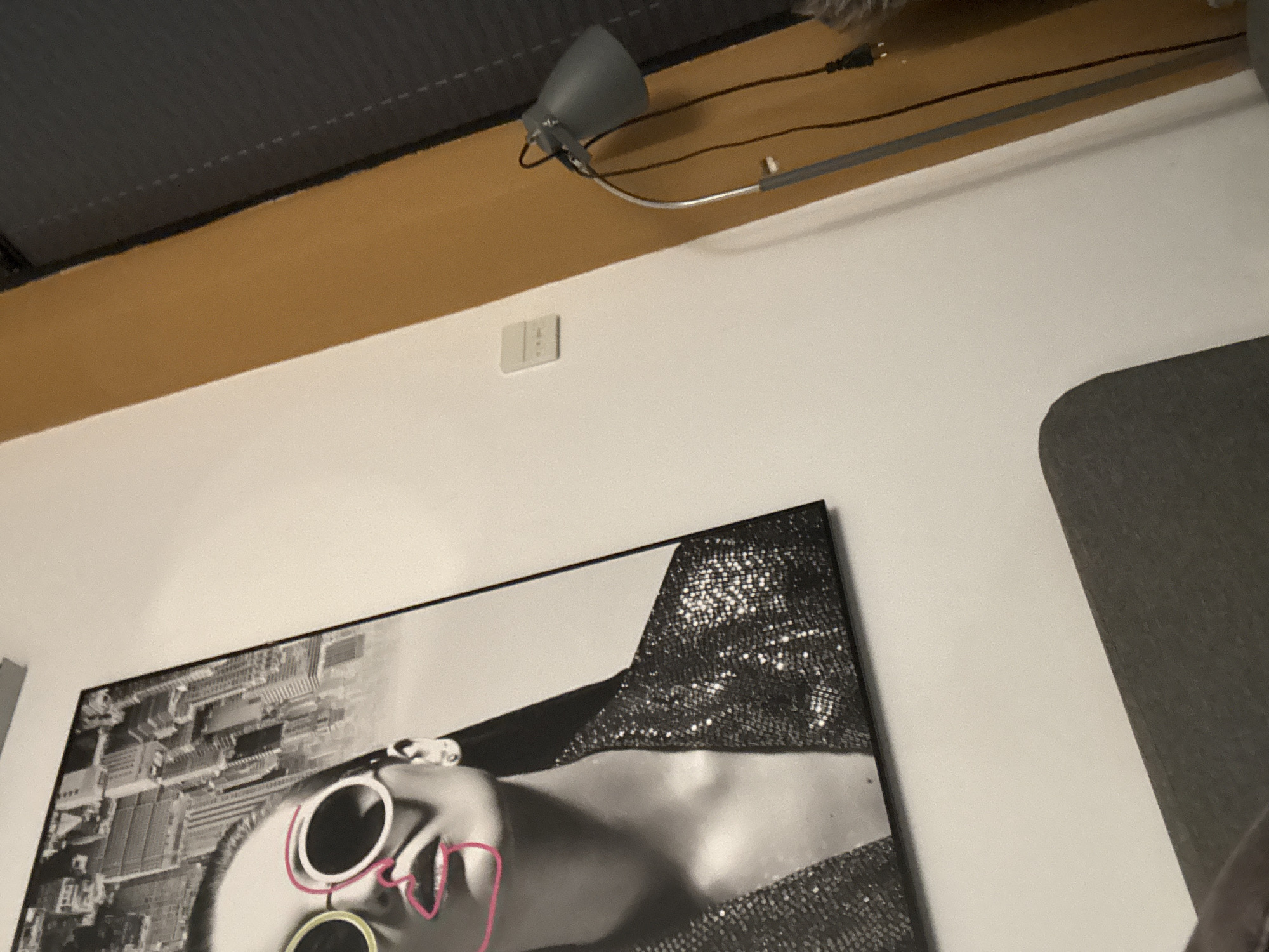 The image shows a close-up of a black and white portrait artwork of a woman with bold pink and yellow sunglasses. The artwork is hanging on a white wall. To the right of the artwork, a portion of a floor lamp with a gray shade is visible, along with its black cord plugged into an outlet. The wall transitions to a mustard yellow color, and further to the right, a dark gray blind is partially visible. In the foreground, a dark gray upholstered piece of furniture, likely a sofa or chair, is visible. The scene appears to be indoors, possibly in a living room or hallway. The location in the city is unknown.