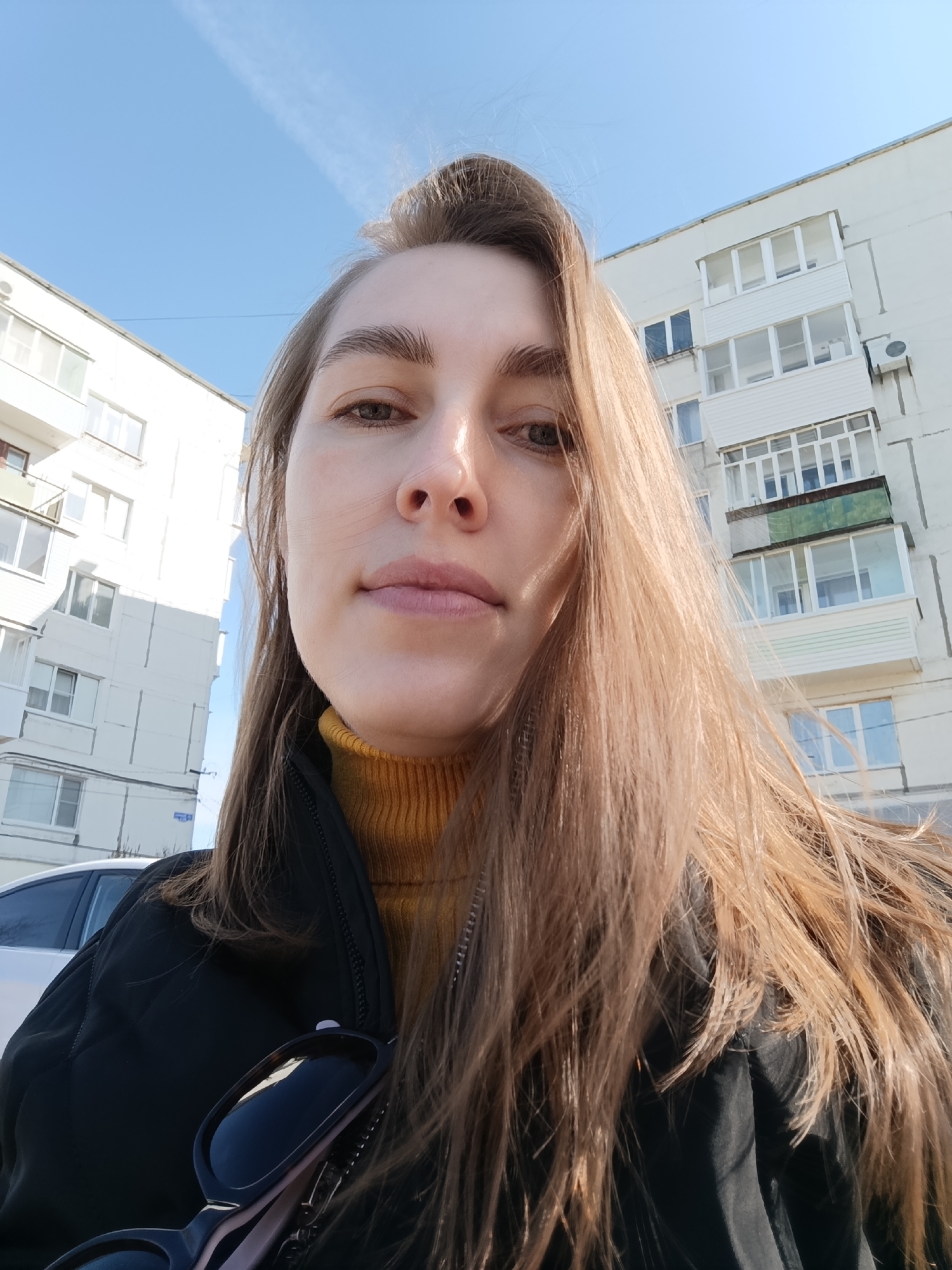 A woman stands outside, facing the camera. She has fair skin, light brown hair, and is wearing a black jacket over a yellow turtleneck sweater. Sunglasses are attached to the front of her jacket. Behind her are residential buildings, indicative of an urban setting. The buildings appear to be white apartment blocks common in Russia. A white car is partially visible on the lower left. The clear blue sky suggests it is daytime. The location is inferred to be Chaikovsky, Russia, based on the request.