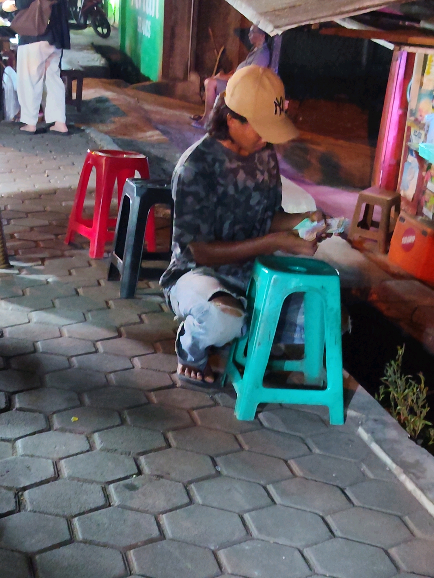 A person wearing a camouflage-patterned shirt and a tan baseball cap is sitting on a teal plastic stool, counting money. The person is also wearing light-colored pants and flip-flops. The scene takes place on a paved area with hexagonal gray stones, suggesting an outdoor market or street setting, possibly in Purwakarta, Indonesia, given the context. In the background, there are other plastic stools, one red and one black, and what appears to be a small stall with colorful items and signage. The lighting suggests it is evening or night.