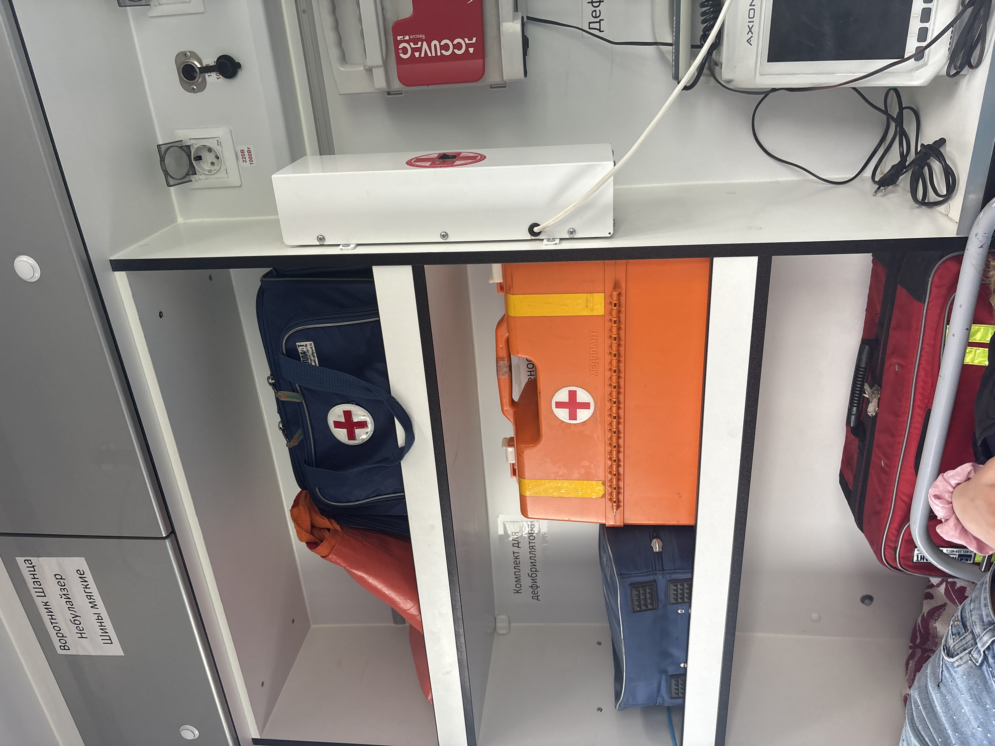 The image shows the interior of an ambulance, with medical supplies neatly organized on shelves. On the top shelf, there is a blue medical bag with a red cross emblem, and to its left, an orange tubular object. A white sticker on the cabinet above reads "Воротник Шанца Небулайзер Шины мягкие" which translates to "Shants collar, Nebulizer, Soft splints."
The middle shelf contains an orange medical case with a red cross, a yellow stripe, and the text "МЕДПЛАНТ" printed on it. To the left of this case is a blue bag with metal stud details. A white sticker on the shelf above reads "Комплект для дефибриллятора" which translates to "Defibrillator kit."
The bottom shelf holds a red bag with black accents, and to the left, a person's legs are visible, wearing blue jeans with a speckled pattern.
On the right side of the ambulance interior, a white device with a red cross emblem is mounted on the wall. Below it, a red package labeled "ACCUVAC Rescue" is visible. Further down, a monitor screen with the brand name "AXION" is present, connected by cables. Electrical outlets and other medical equipment are also visible. The image appears to have been taken in Pesochny, Russia, as indicated by the context provided.