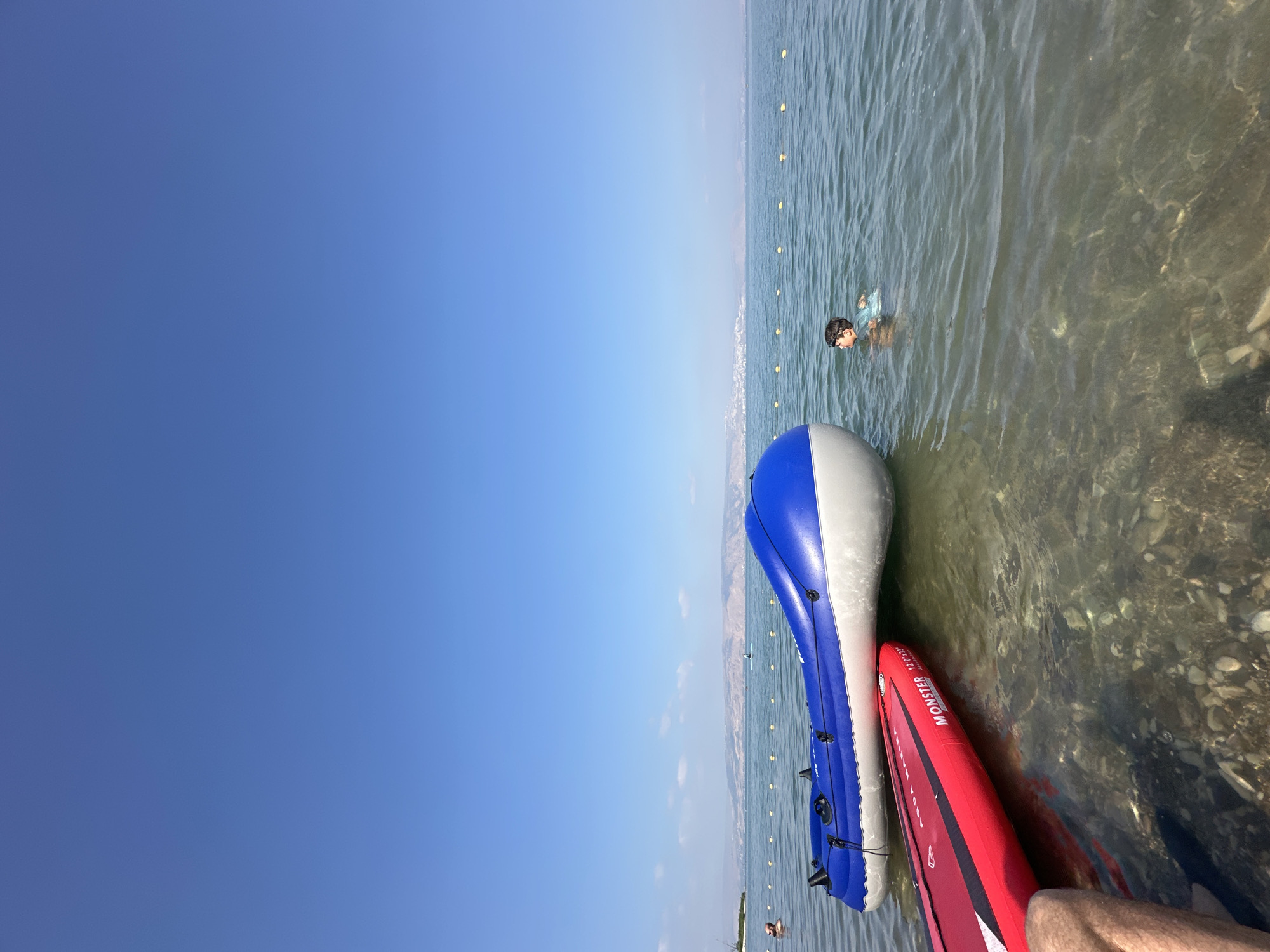 In Tiberias, Israel, on a clear day with a bright blue sky, a red paddleboard branded "MONSTER" is positioned near an inflatable blue and white raft in the shallow, clear water of the Sea of Galilee. A young boy wearing a light blue t-shirt is swimming in the water to the right of the raft, looking towards the distant shore. The seabed is visible beneath the shallow water, covered with stones and pebbles. Yellow buoys mark a boundary in the water, and a hazy, mountainous landscape with buildings can be seen on the opposite shore.