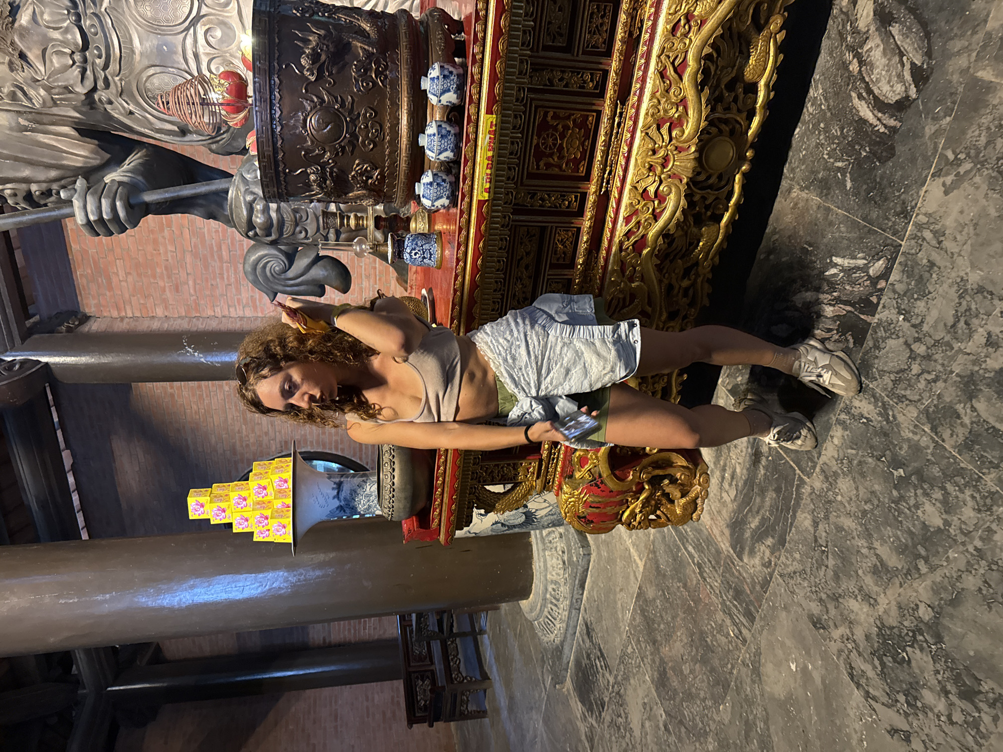 In Tây Hoa Lư Ward, Vietnam, a woman with curly brown hair stands in what appears to be a temple or shrine. She is wearing a light-colored tank top, green shorts, and white athletic shoes. A light-colored garment is tied around her waist. She is looking to her right and holding a yellow object in her right hand, near her face. In front of her is an ornate, red and gold altar with various objects. These include a large, dark bronze urn decorated with dragons, three small blue and white ceramic bowls, a small blue and white vase, and a stack of yellow boxes with pink floral designs arranged in a pyramid on top of a large blue and white vase. Behind the altar, a colossal statue of a warrior figure is visible, holding a staff. The floor is made of large, dark gray stone tiles with lighter gray veining. In the background, a brick wall and large wooden columns are present, along with a wooden bench. The lighting suggests an indoor environment, possibly with artificial lights.